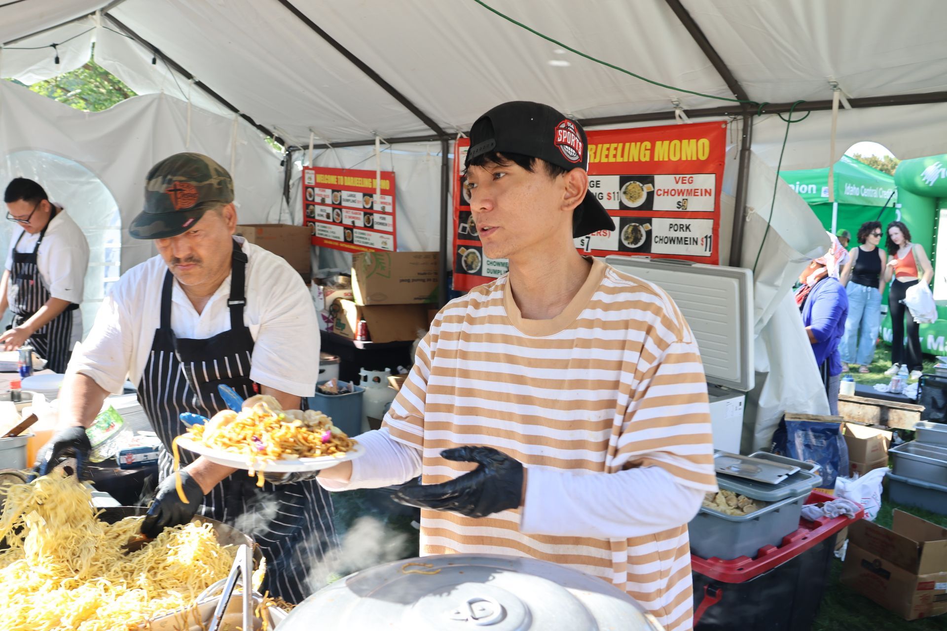 People preparing and serving food at an outdoor food stall. One holds a plate, another cooks noodles.