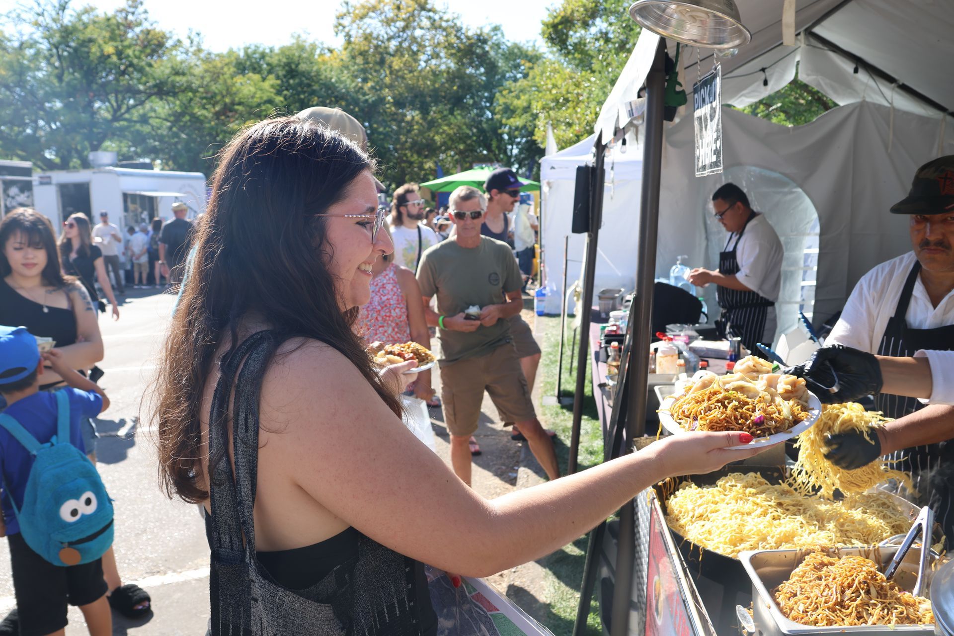 Woman buying food from a vendor at an outdoor food fair, smiling. Others in the background.