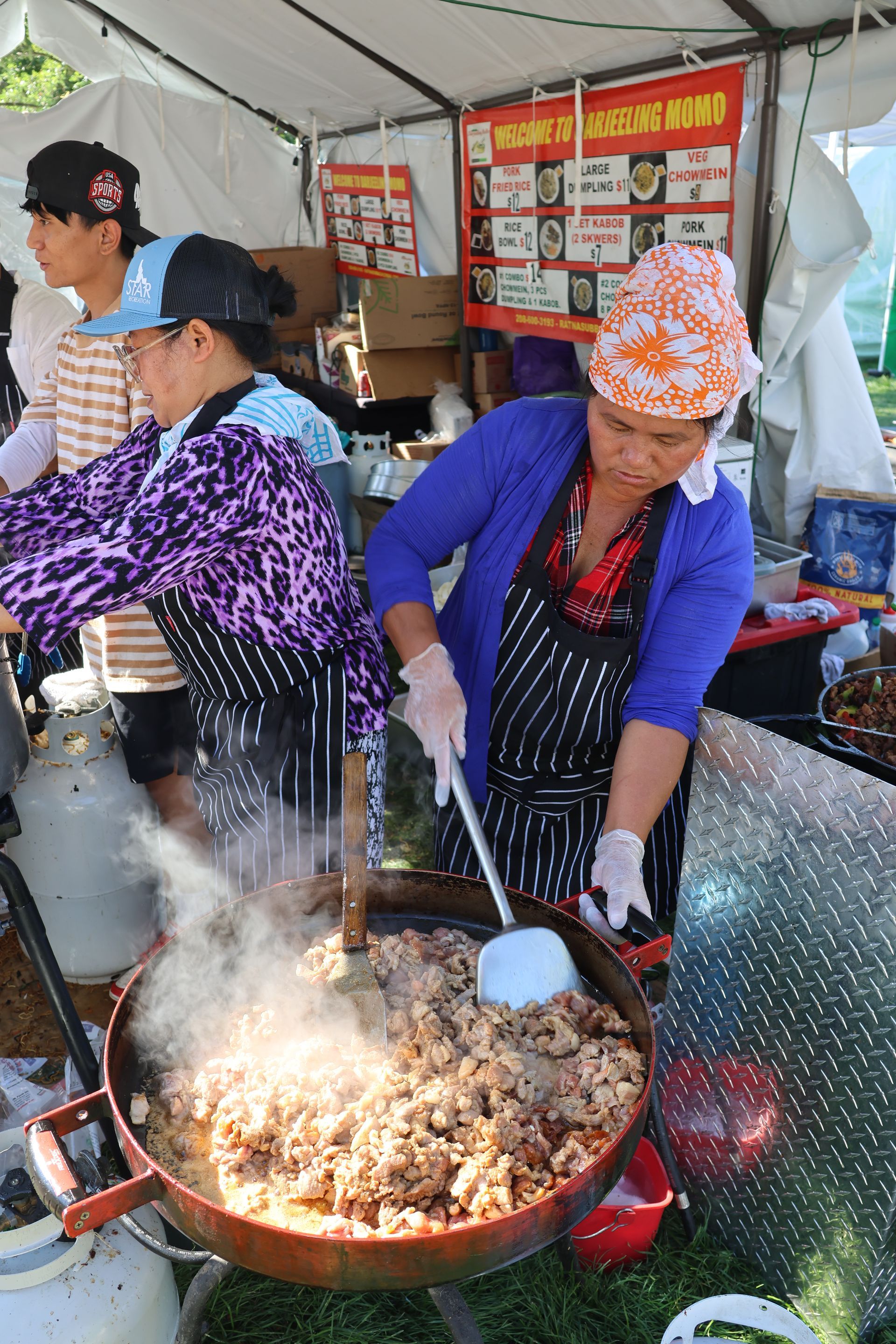 People cooking food in a large pan at a food stall. Woman stirs food, steam rising.