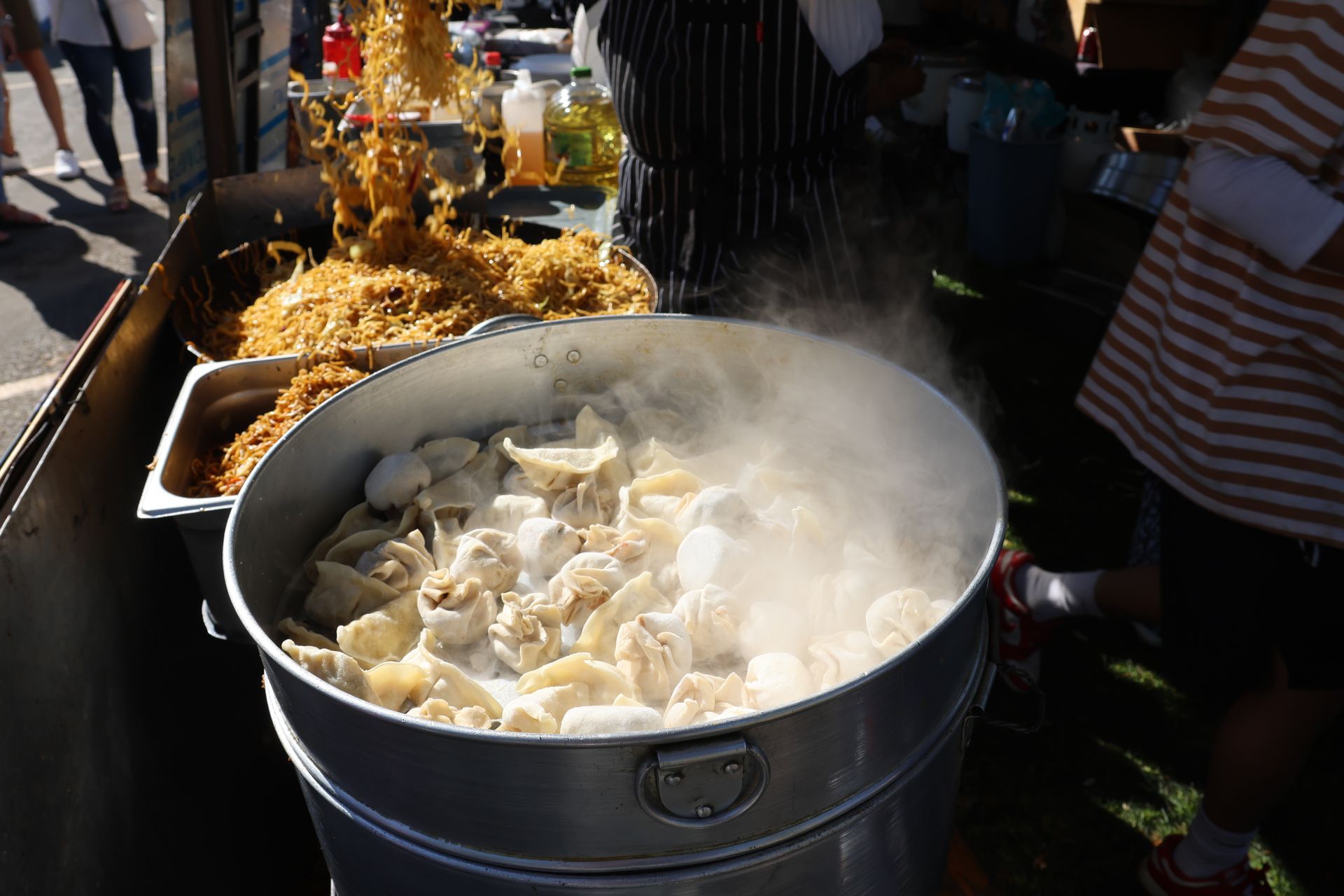 Steaming dumplings in a metal pot at an outdoor food stall, with noodles and a chef visible.