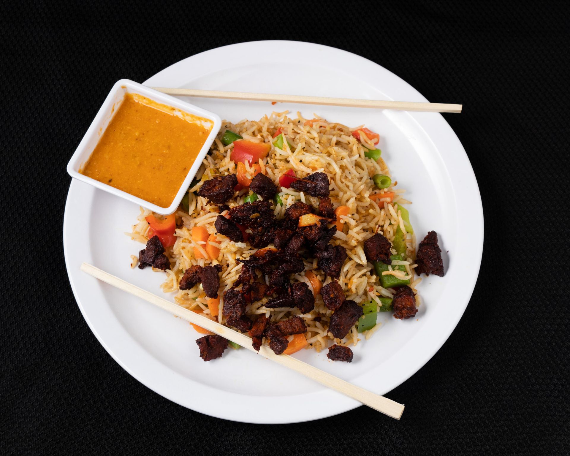 Fried rice with vegetables and meat on a white plate with a small bowl of sauce and chopsticks.