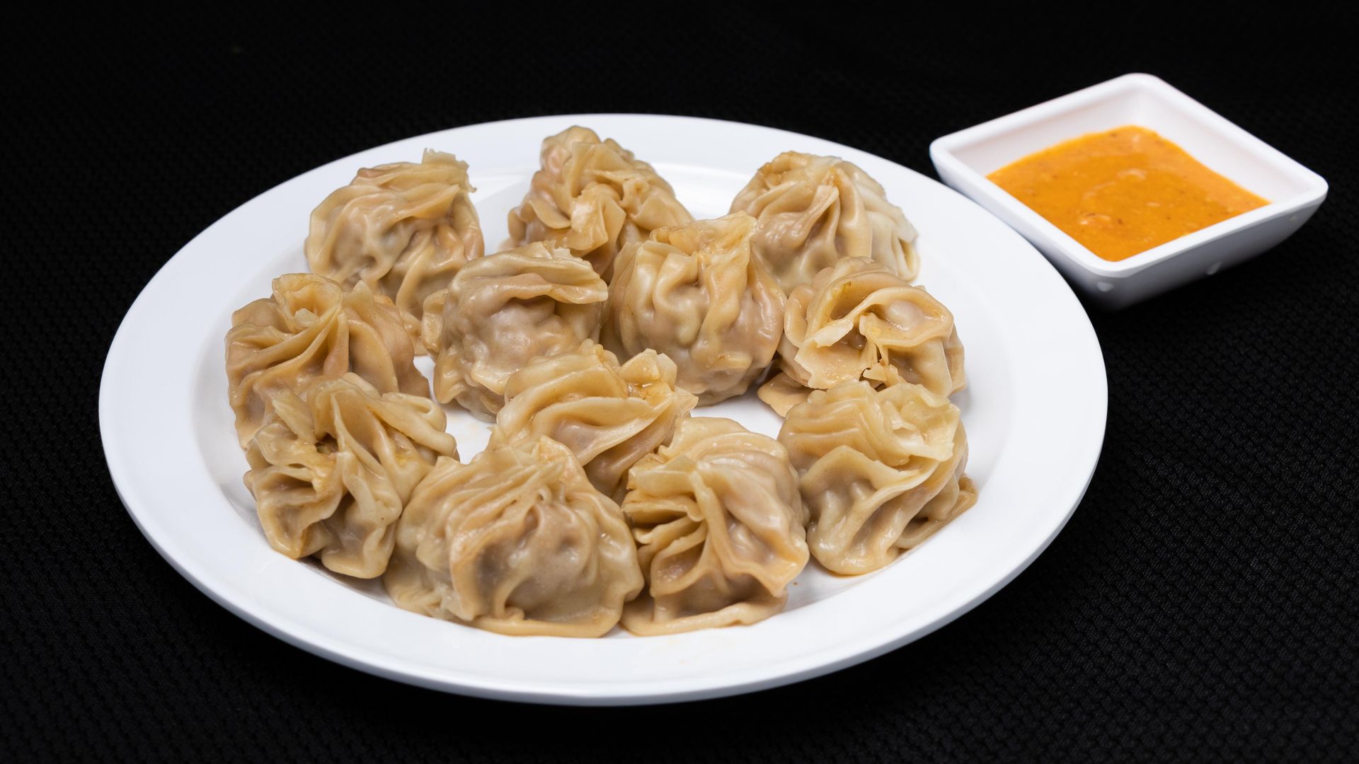 Steamed momos on a white plate with orange dipping sauce on a black background.
