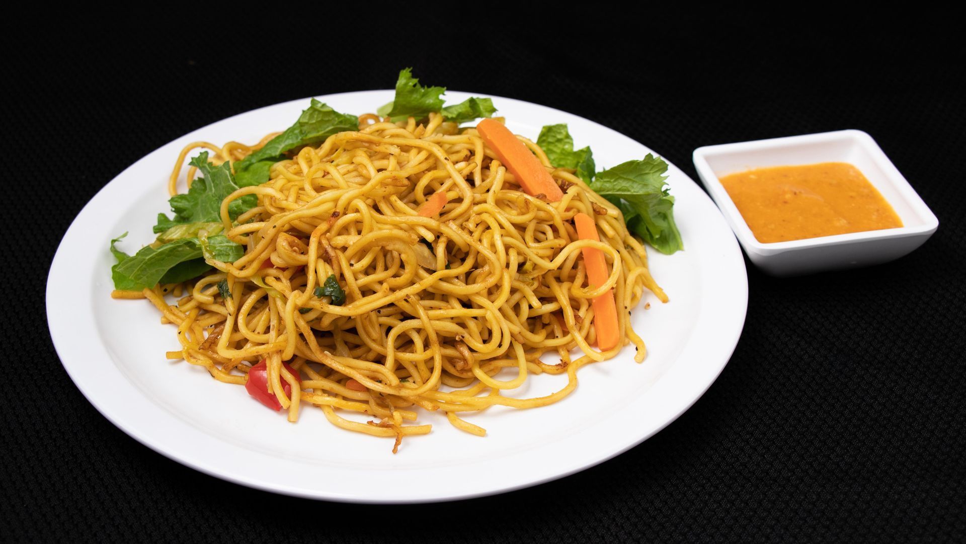 Noodle dish with carrots and greens on a white plate, with a side of orange sauce, set against a black background.