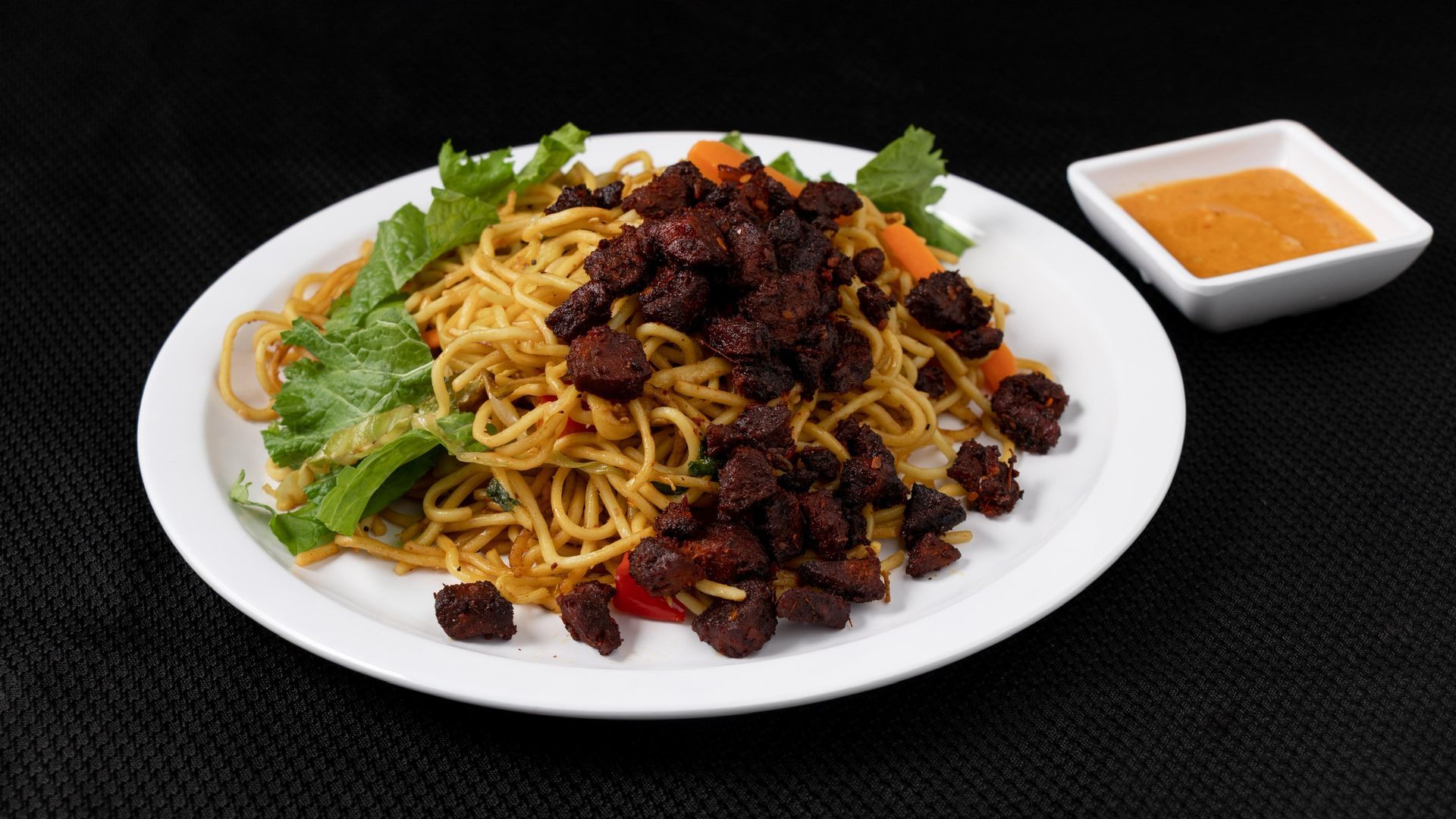 Noodles topped with meat and greens on a plate, with a sauce cup alongside.