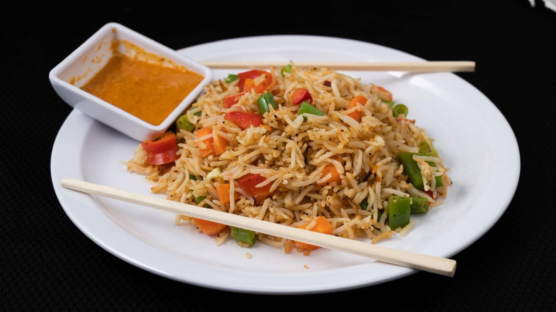 Fried rice with vegetables on a white plate, served with dipping sauce and chopsticks.