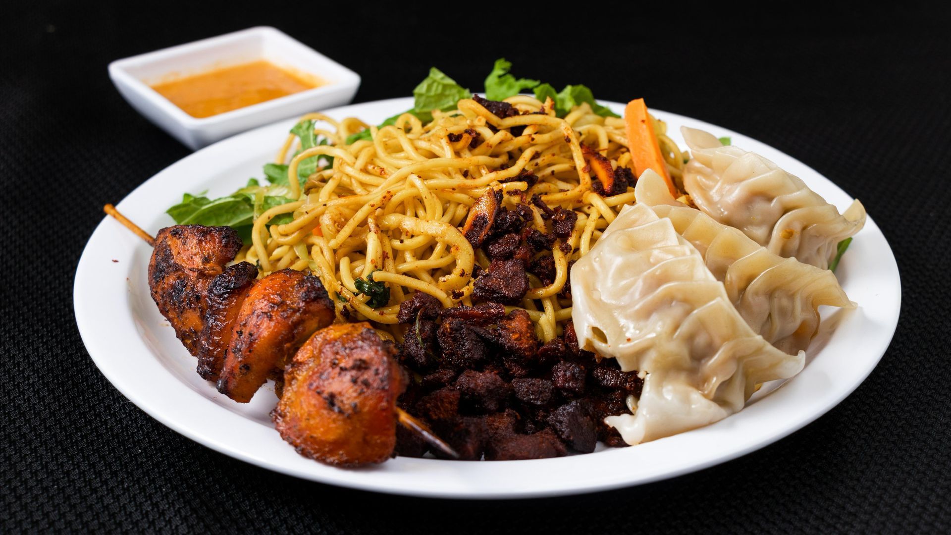Plate of food with noodles, dumplings, skewers, and a dipping sauce.
