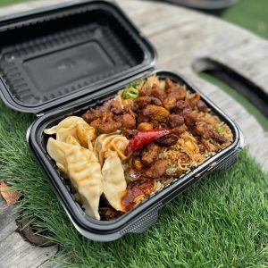 Black takeout container with fried rice, chicken, dumplings on a green surface.