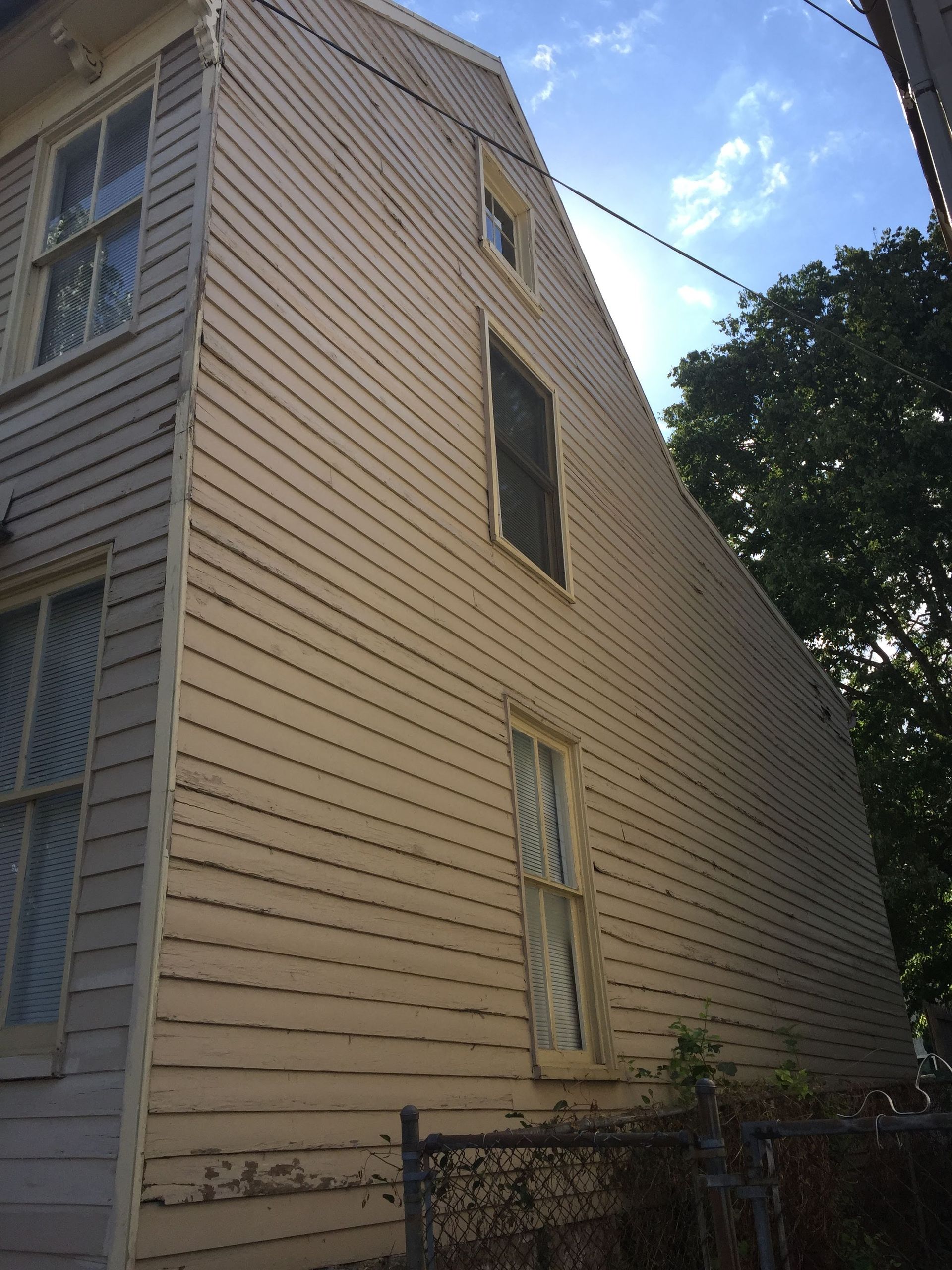 Side of a weathered, beige clapboard building with three windows, a screen, and a chain-link fence.