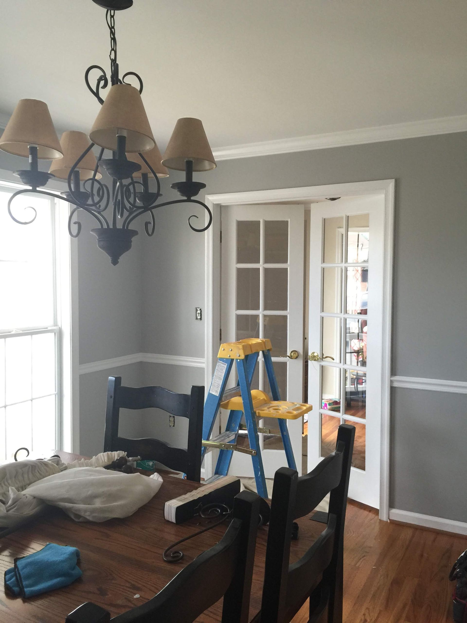 Dining room with chandelier, table, chairs, and French doors. A step ladder stands near the doors. Gray walls and white trim.