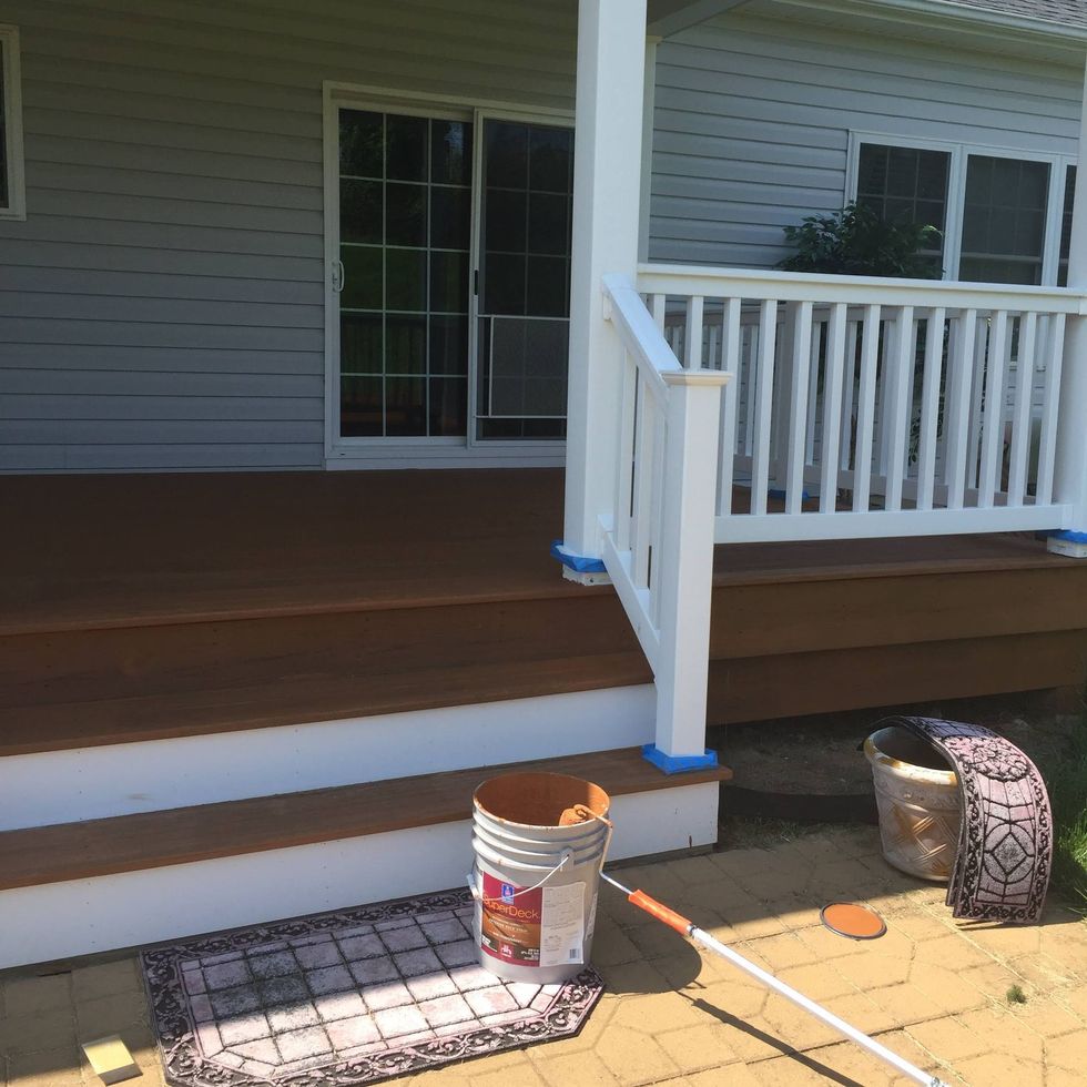 A porch with brown stained steps and white railings. A paint bucket and roller are on the ground.