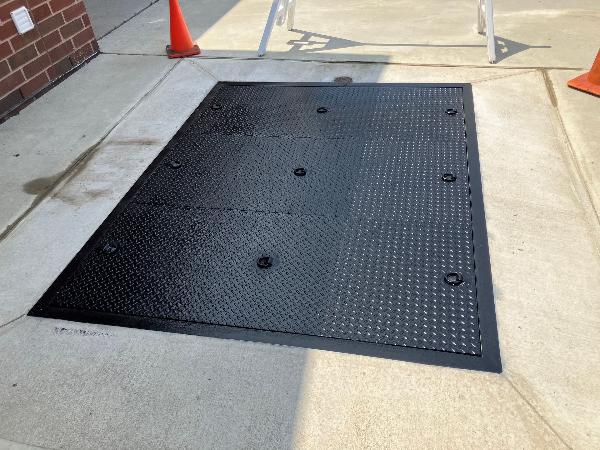 A black manhole cover is sitting on a sidewalk next to a cone.