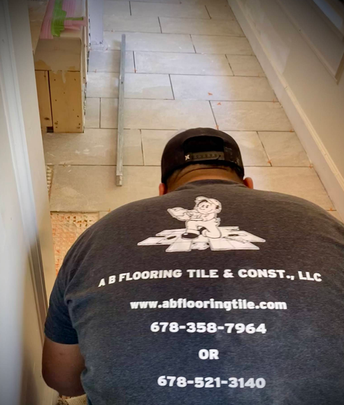 Man installing tile; hallway view. Gray tile, AB Flooring shirt, black cap. Website and phone numbers visible.