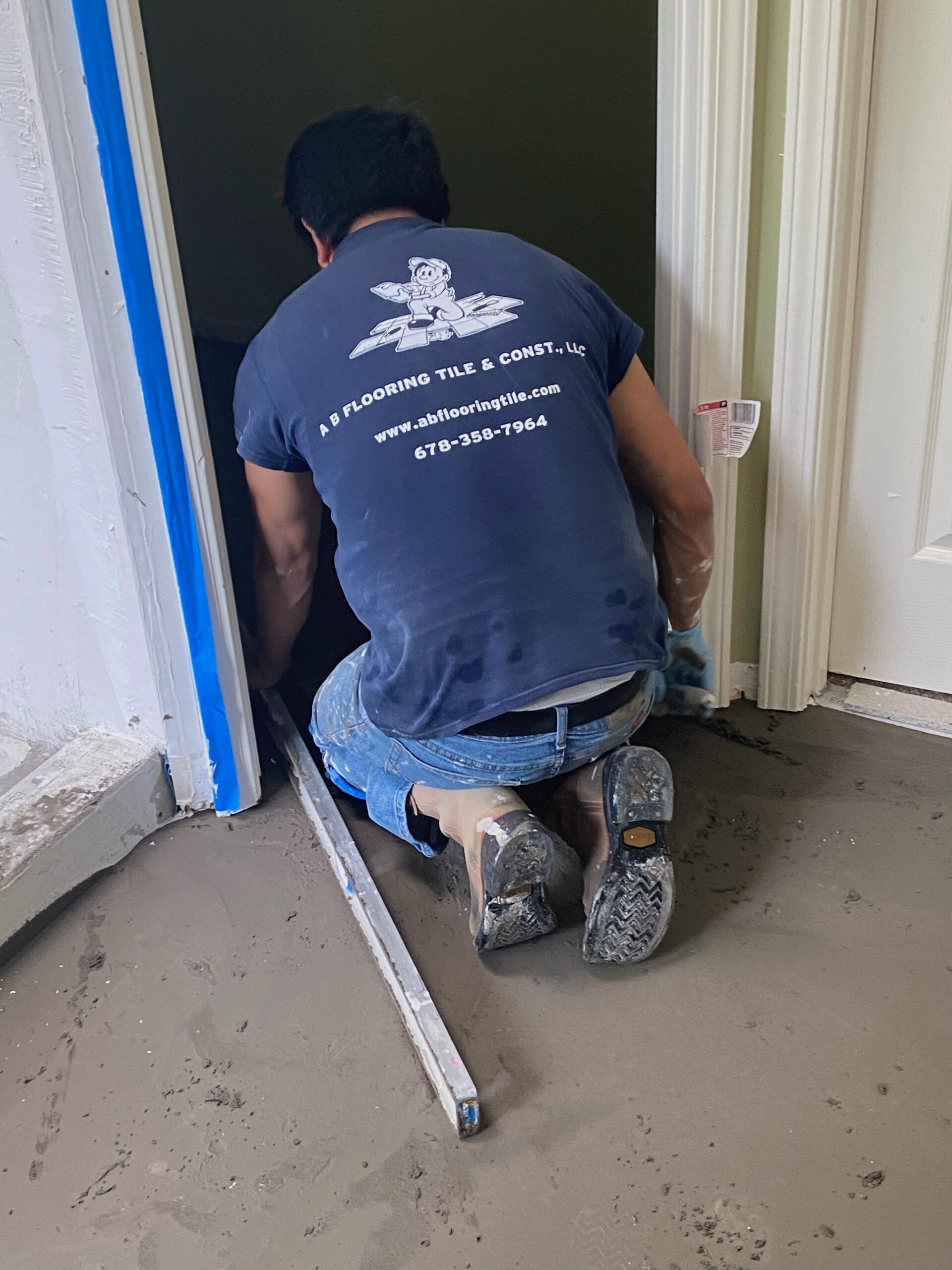 Person on knees, leveling concrete in doorway. Blue shirt, jeans, white door frame.