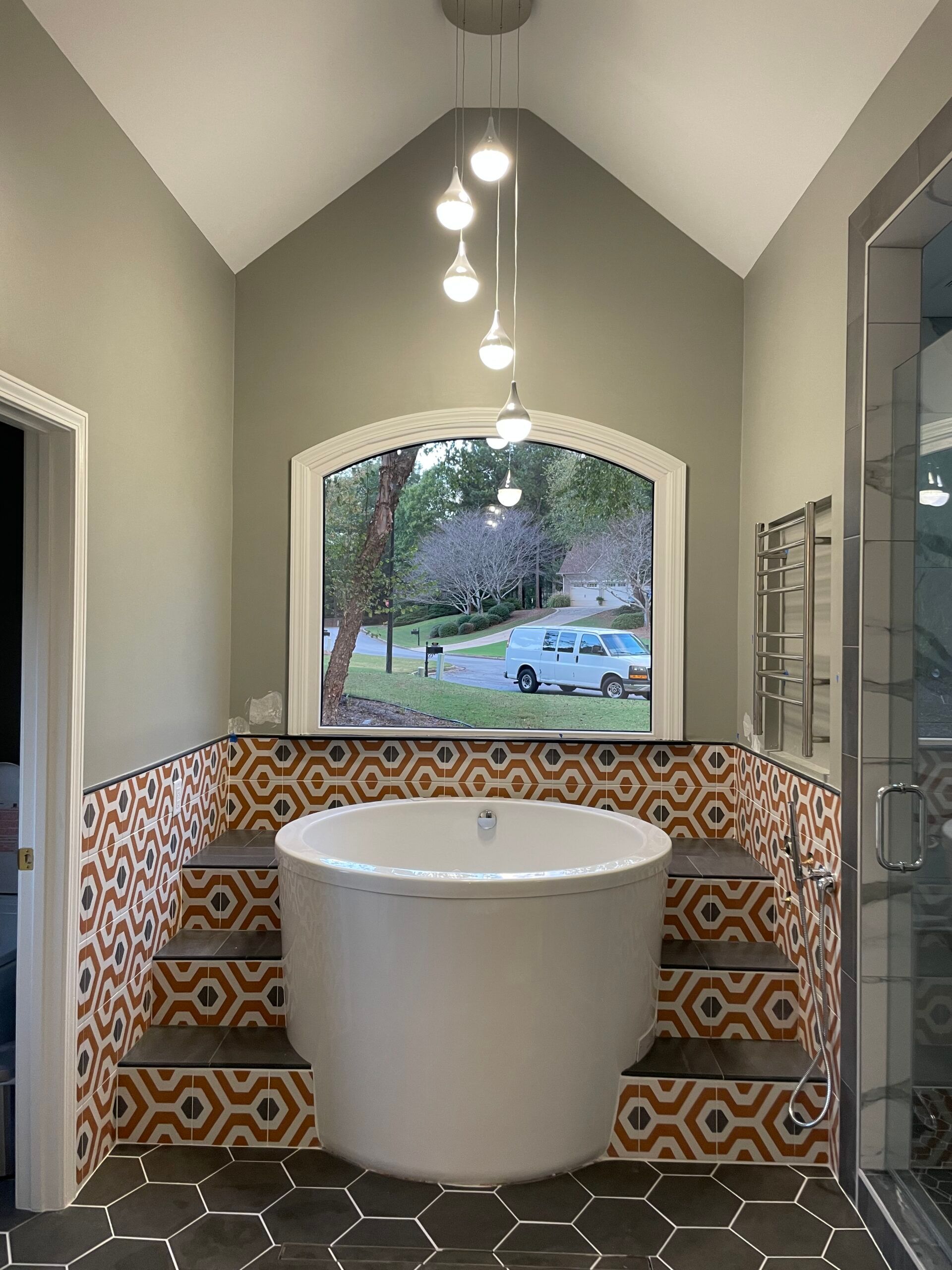 Bathroom with round tub set in tiled steps, arched window above. Neutral walls, dark hexagonal floor tiles.