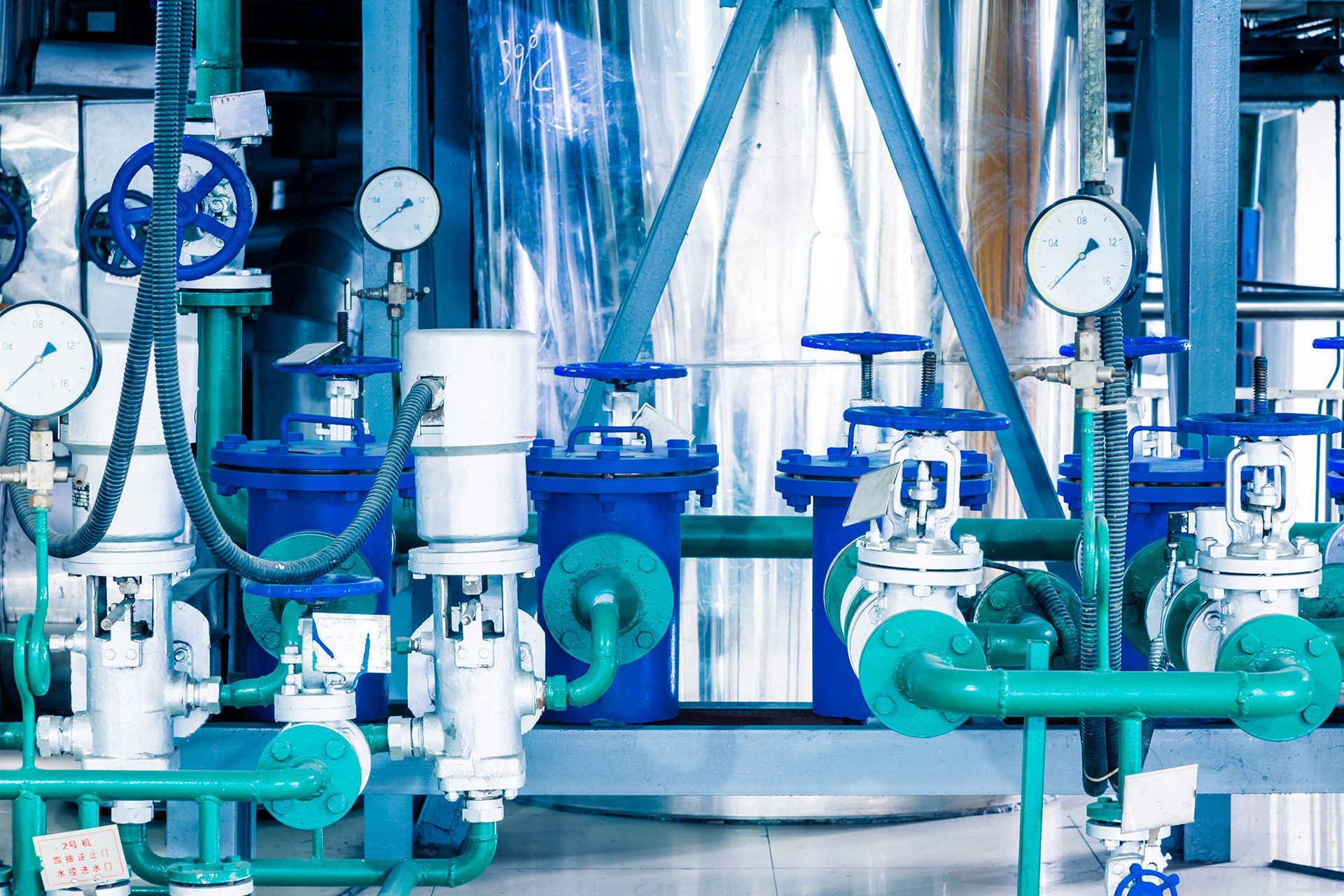 Piping and valves in a factory setting, with blue and green pipes, pressure gauges.