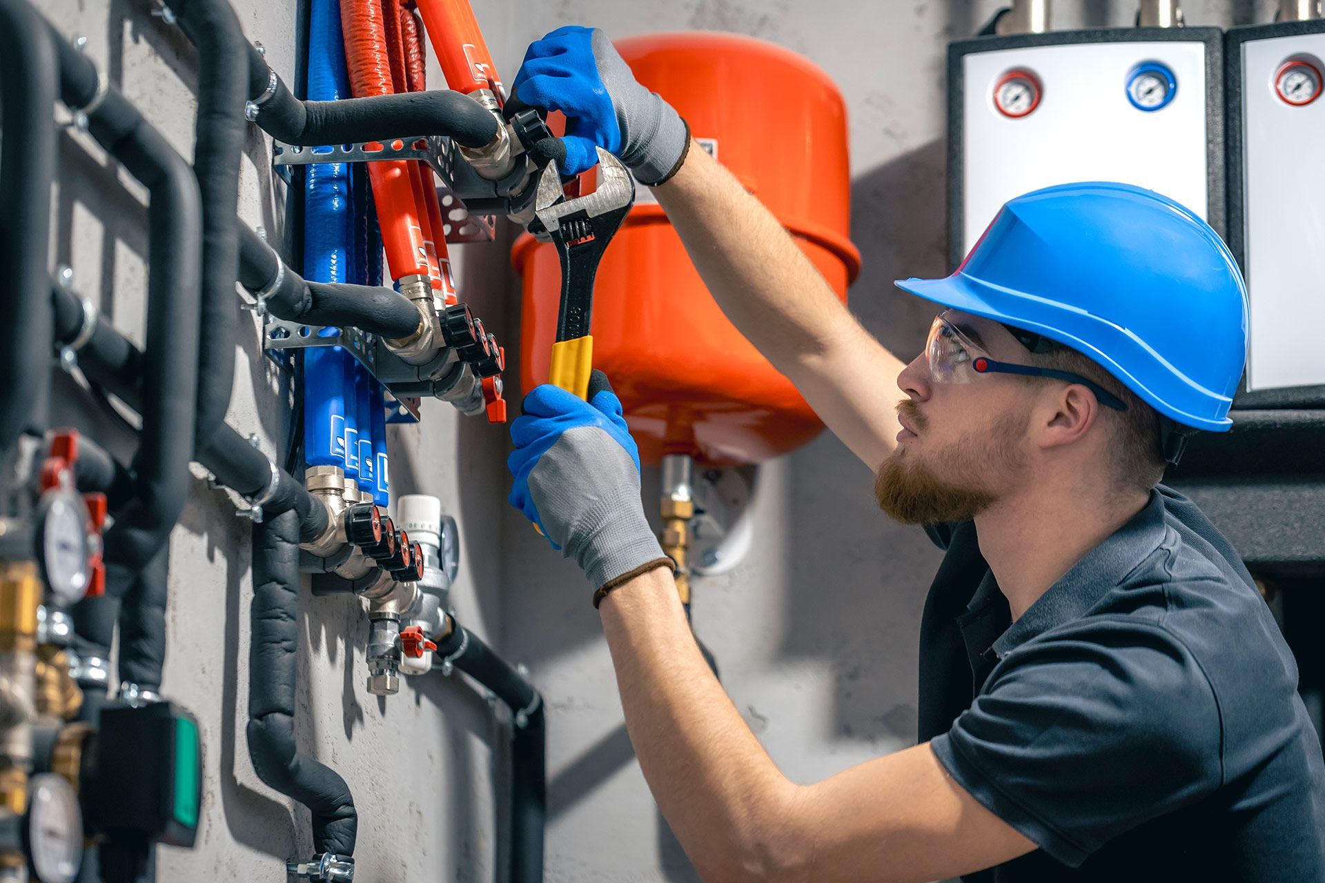 A worker in a blue hard hat and gloves is repairing plumbing with a wrench.