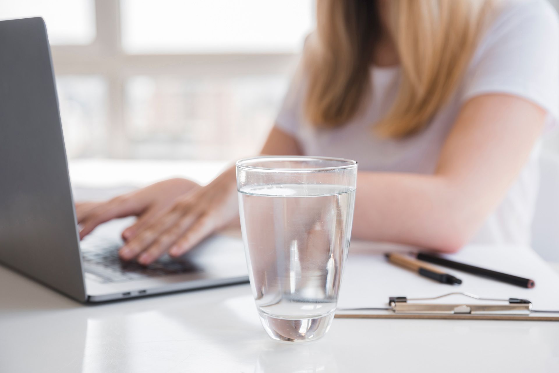 Glass of water on a desk with a laptop and a person typing in the background.