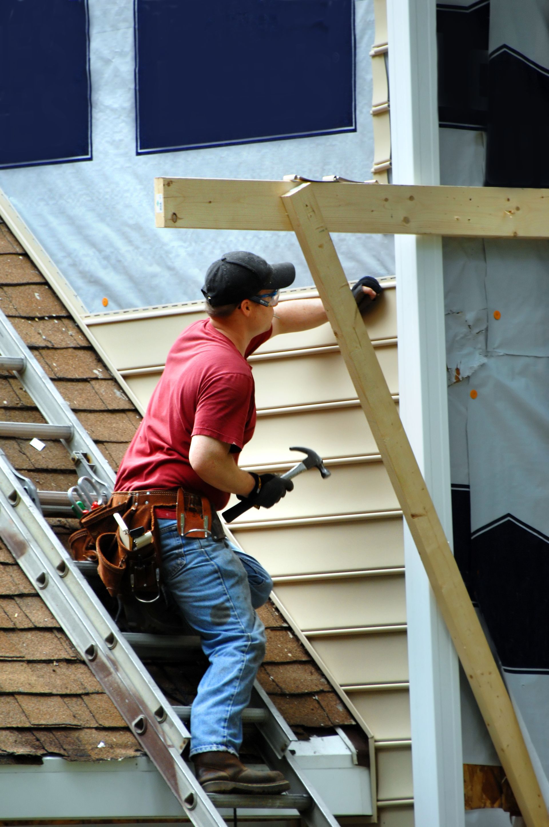 Construction worker on a ladder hammering siding onto a house, wearing a red shirt and tool belt.