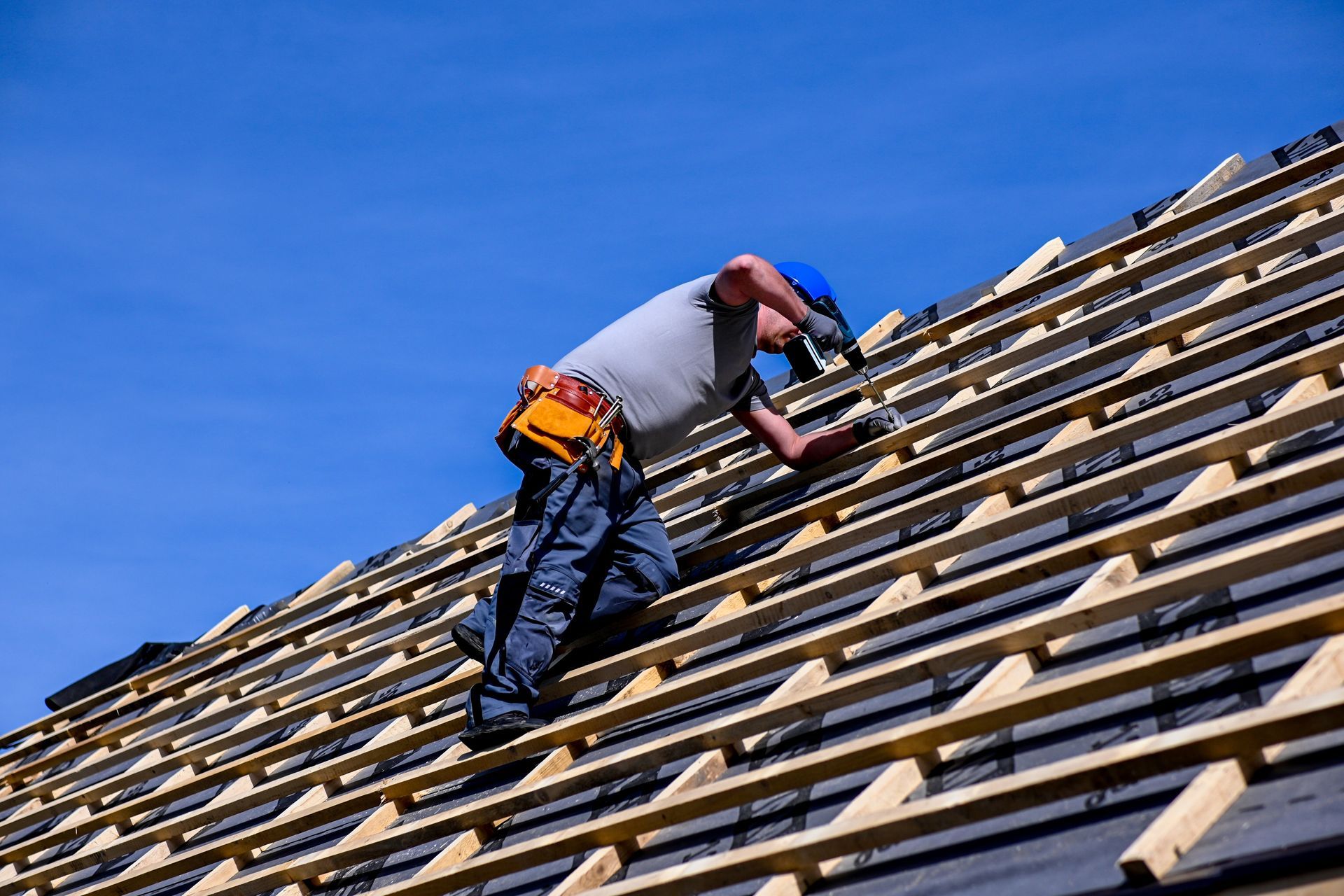 Roofer in blue hard hat installing shingles on a roof under a blue sky.