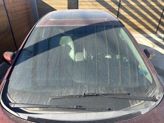 A red car with a dirty windshield is parked in a driveway.