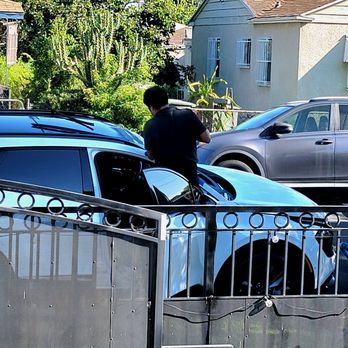 A man is standing next to a blue car in front of a fence.