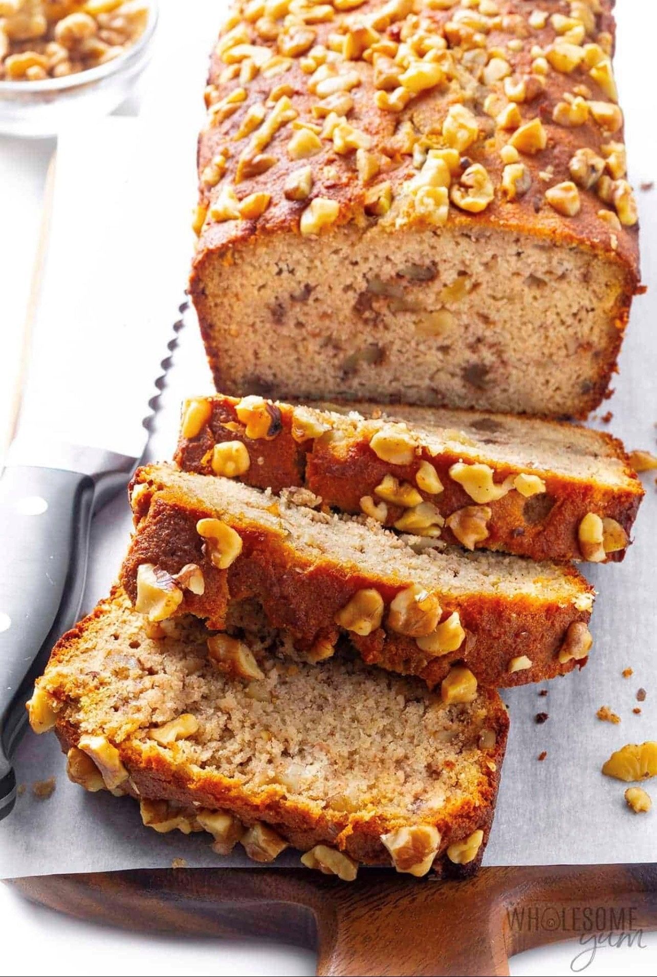 A loaf of banana bread with walnuts on top is sliced on a cutting board.