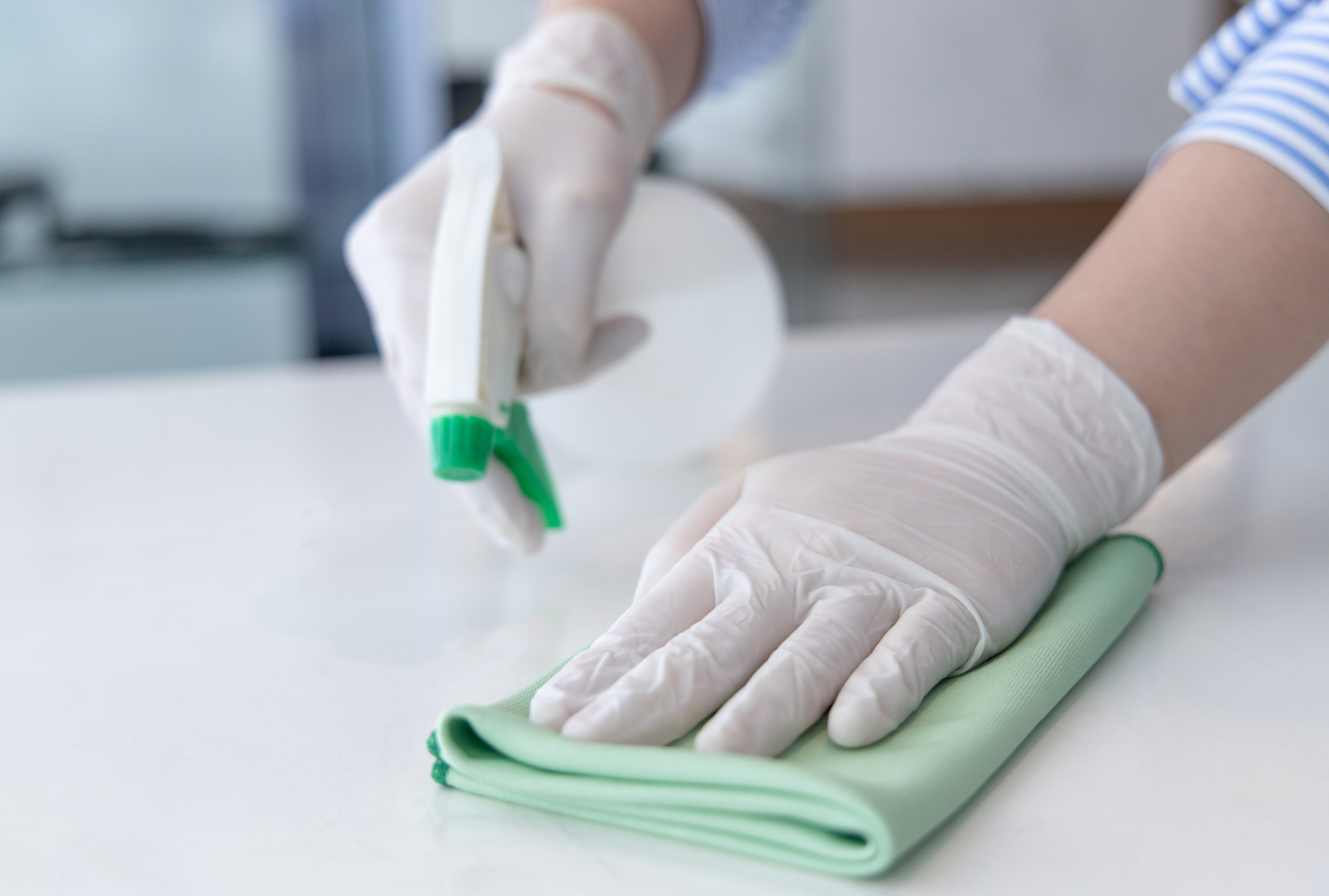 A person wearing gloves is cleaning a table with a cloth and spray bottle.