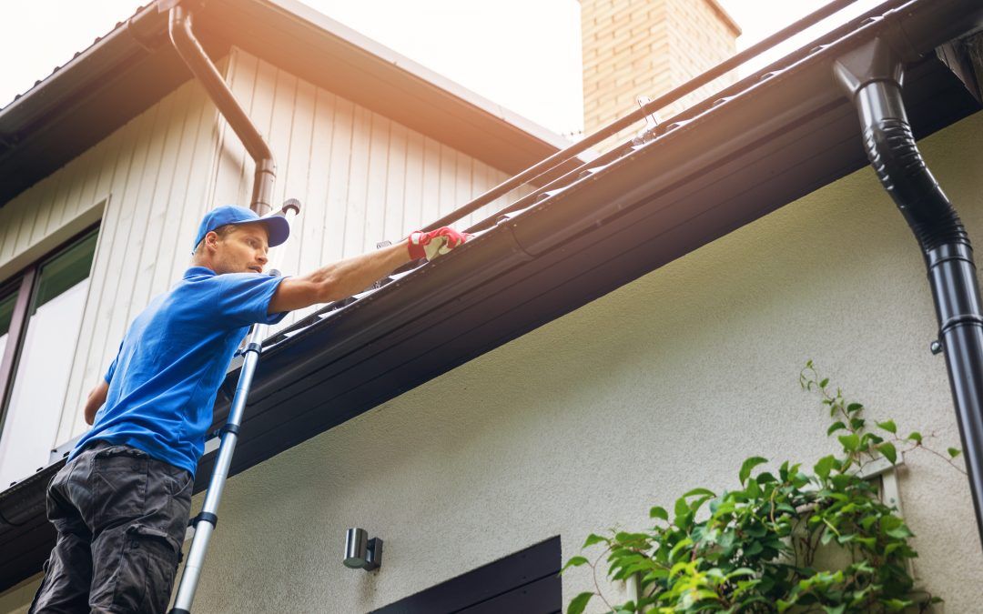 Man on a ladder repairs gutter on a house with brown roof and beige siding during autumn.