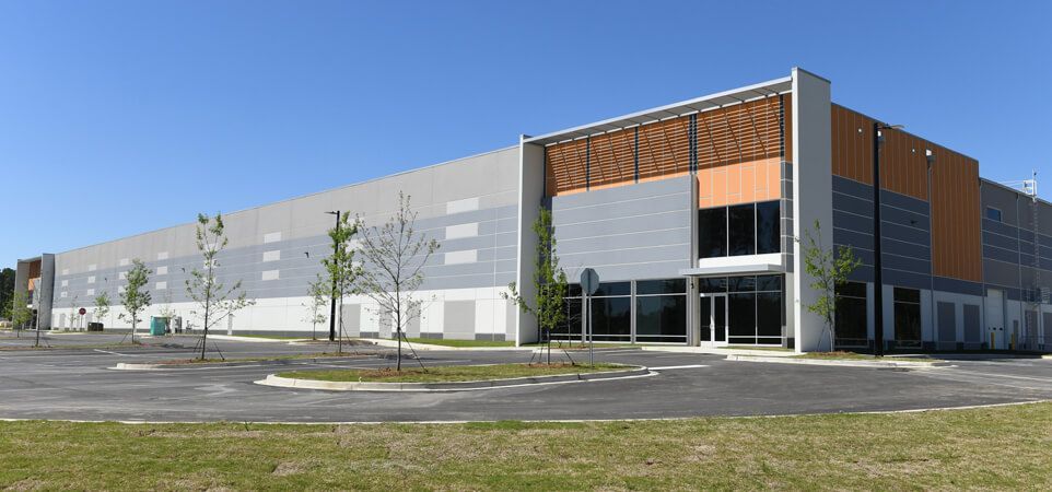 Shopping center with various storefronts under a clear blue sky, cars parked in front.