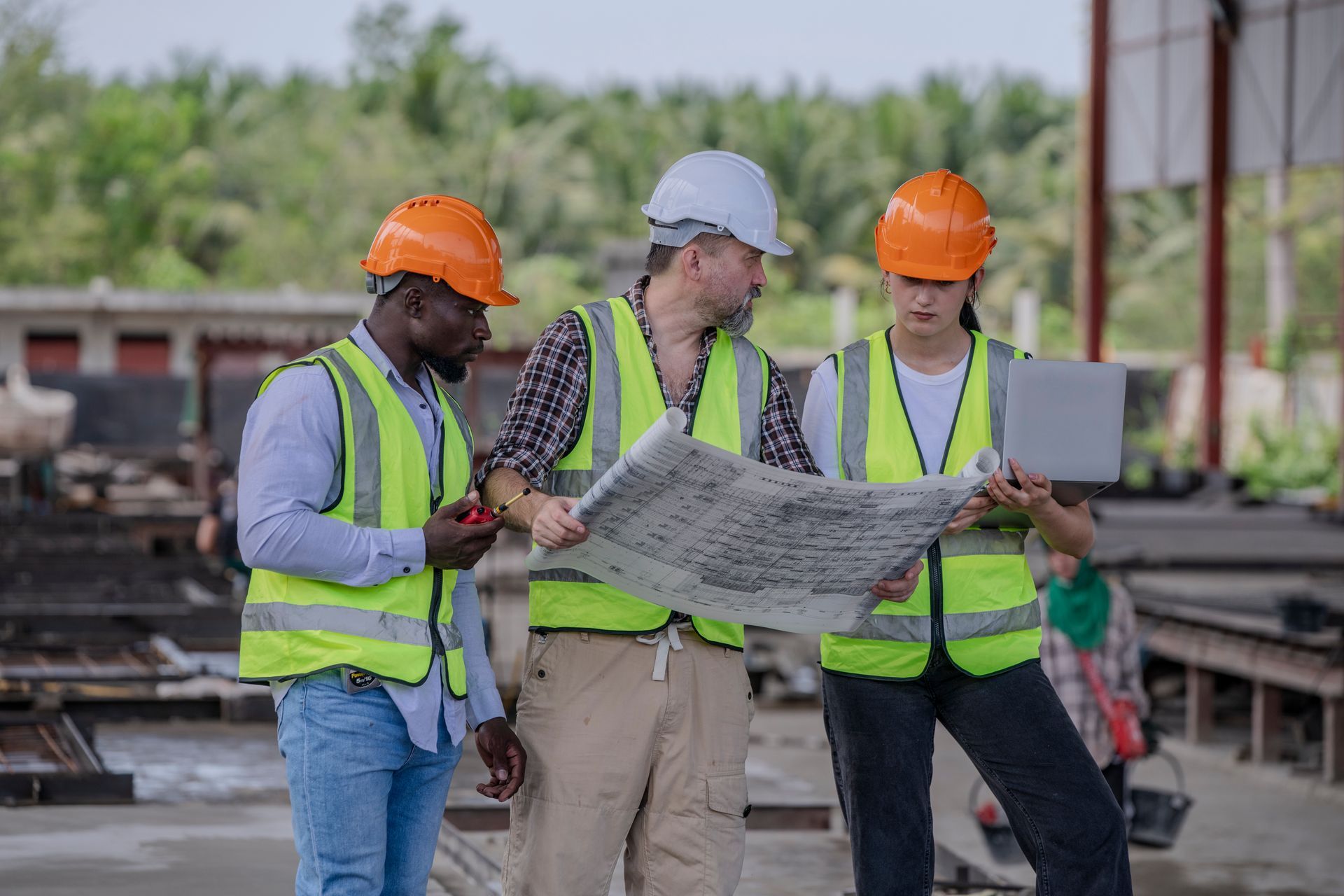Construction worker in orange hardhat uses tablet, team members view plans on rooftop.