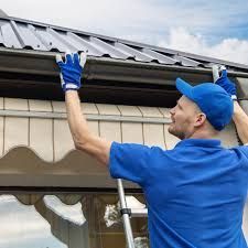 Man on rooftop installing blue roofing, holding tools, rural setting.