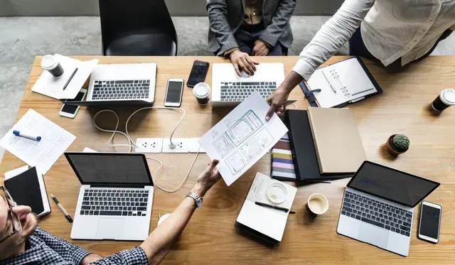A group of people are sitting around a table with laptops and papers.