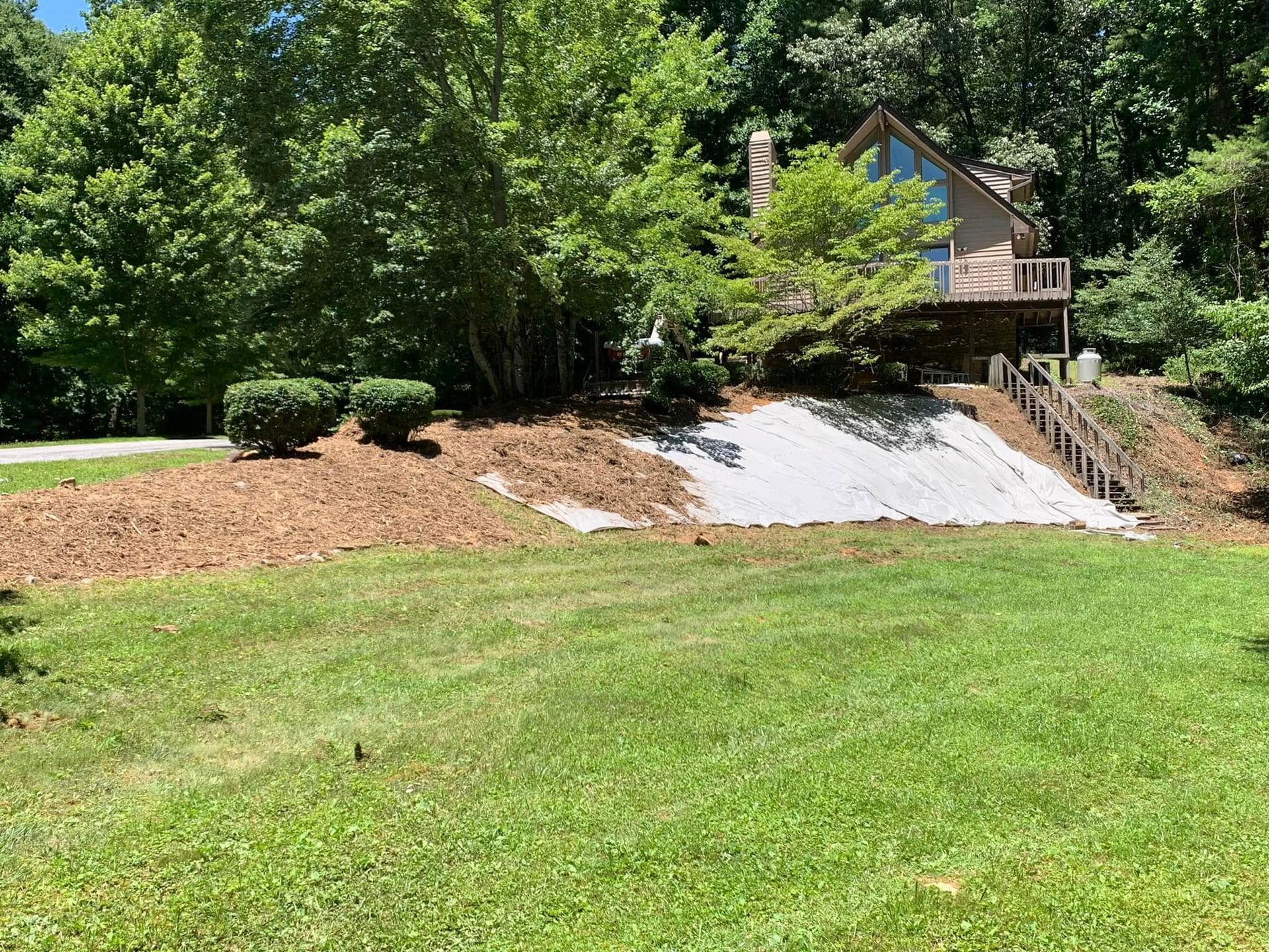 A multi-story wooden house nestled in a lush, green woodland, featuring a mulched slope and an exposed fabric ground cover.
