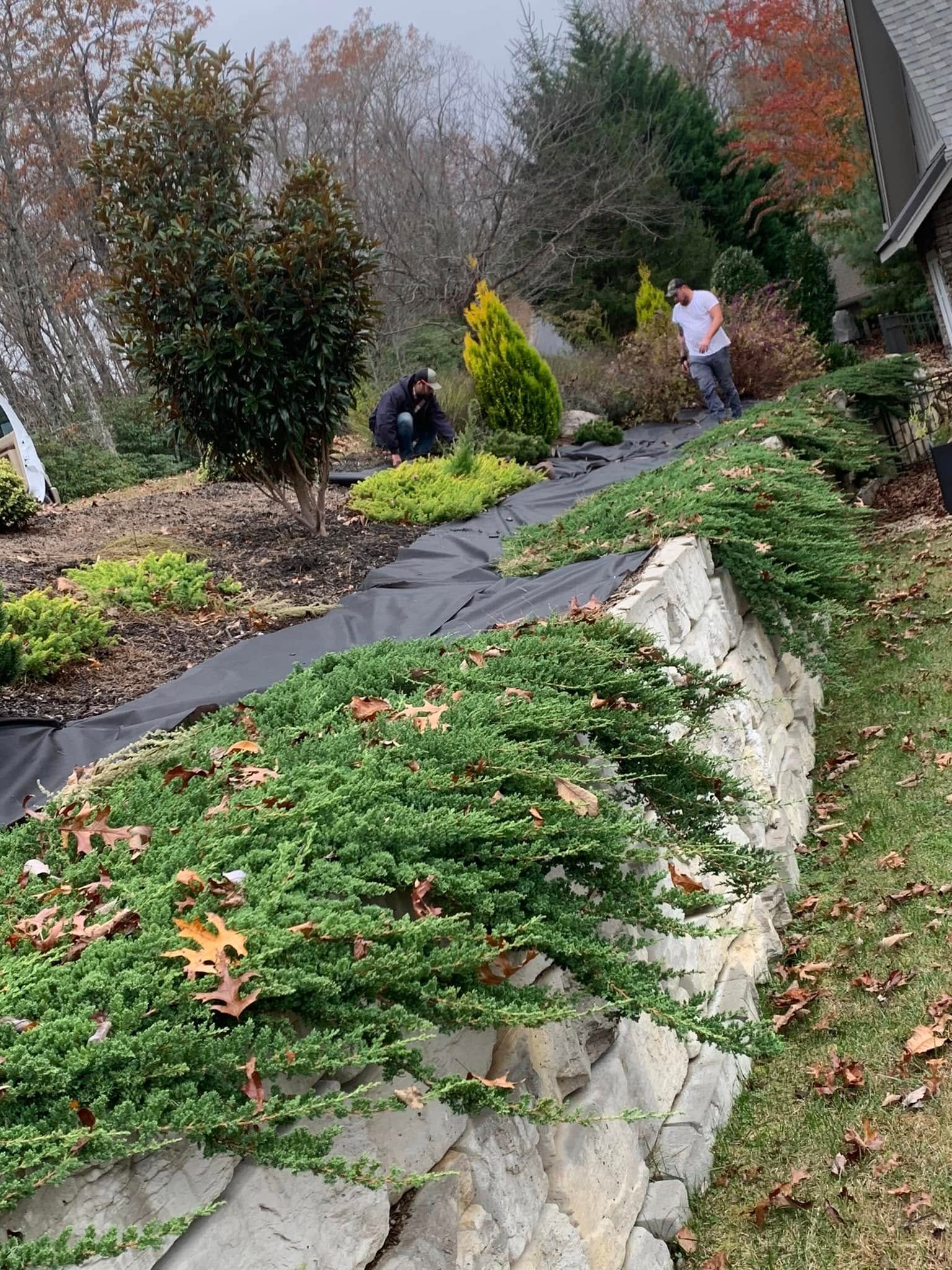 Two people working on landscaping with black garden fabric near a stone retaining wall in an autumnal garden.