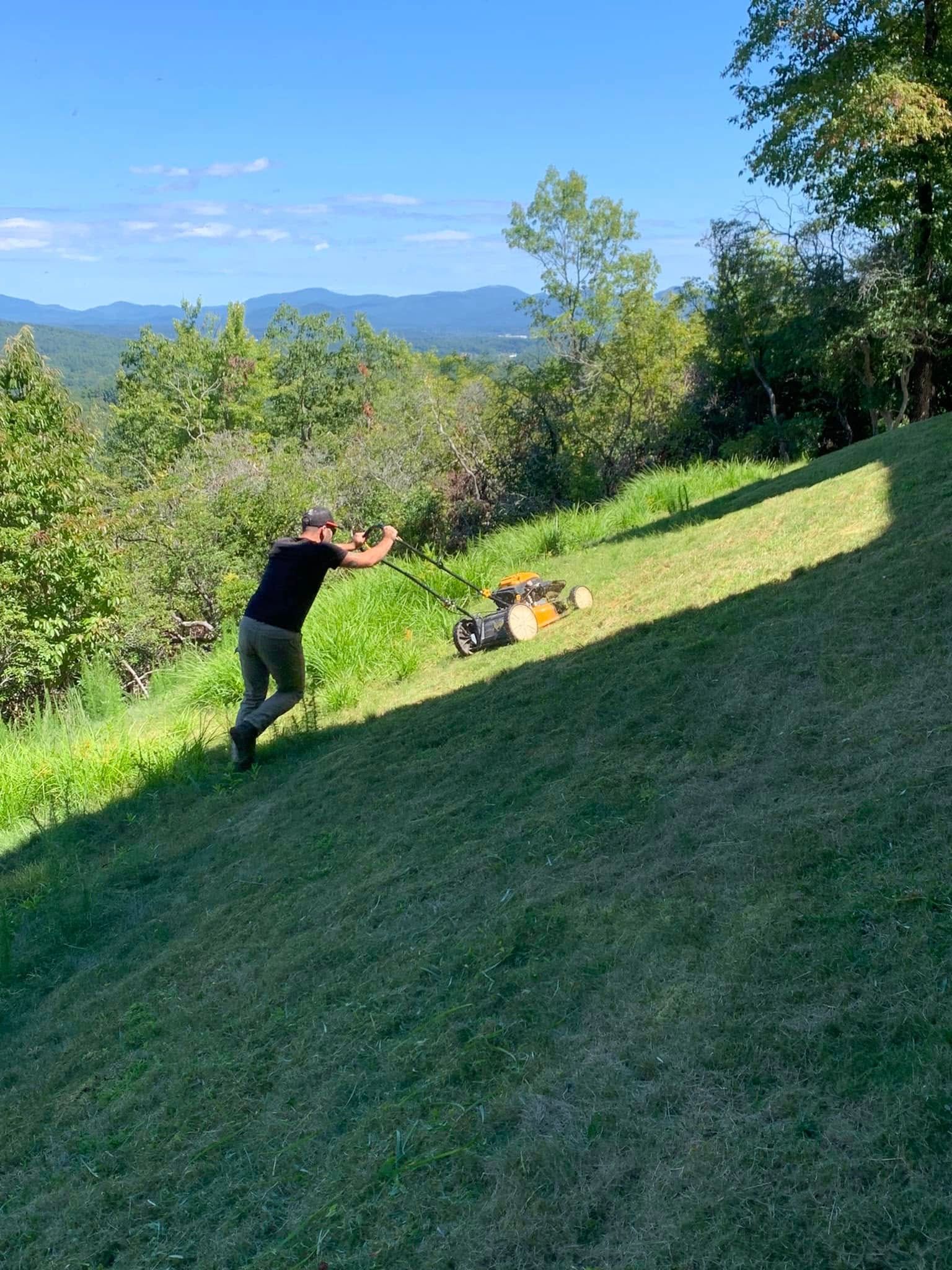 A person in a black shirt and cap pushes a lawn mower up a steep, grassy hillside on a sunny day.
