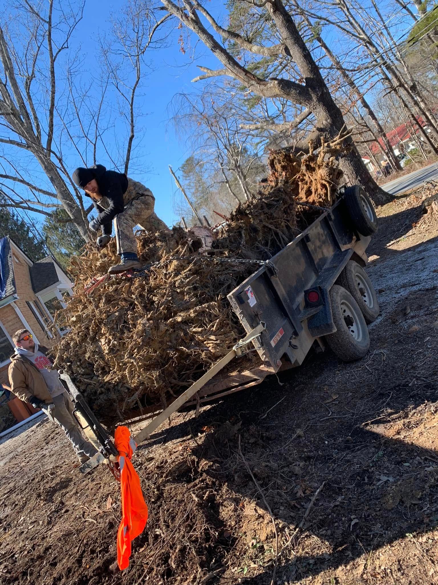 A person stands atop a trailer filled with dry leaves and branches, while another person assists from the ground.