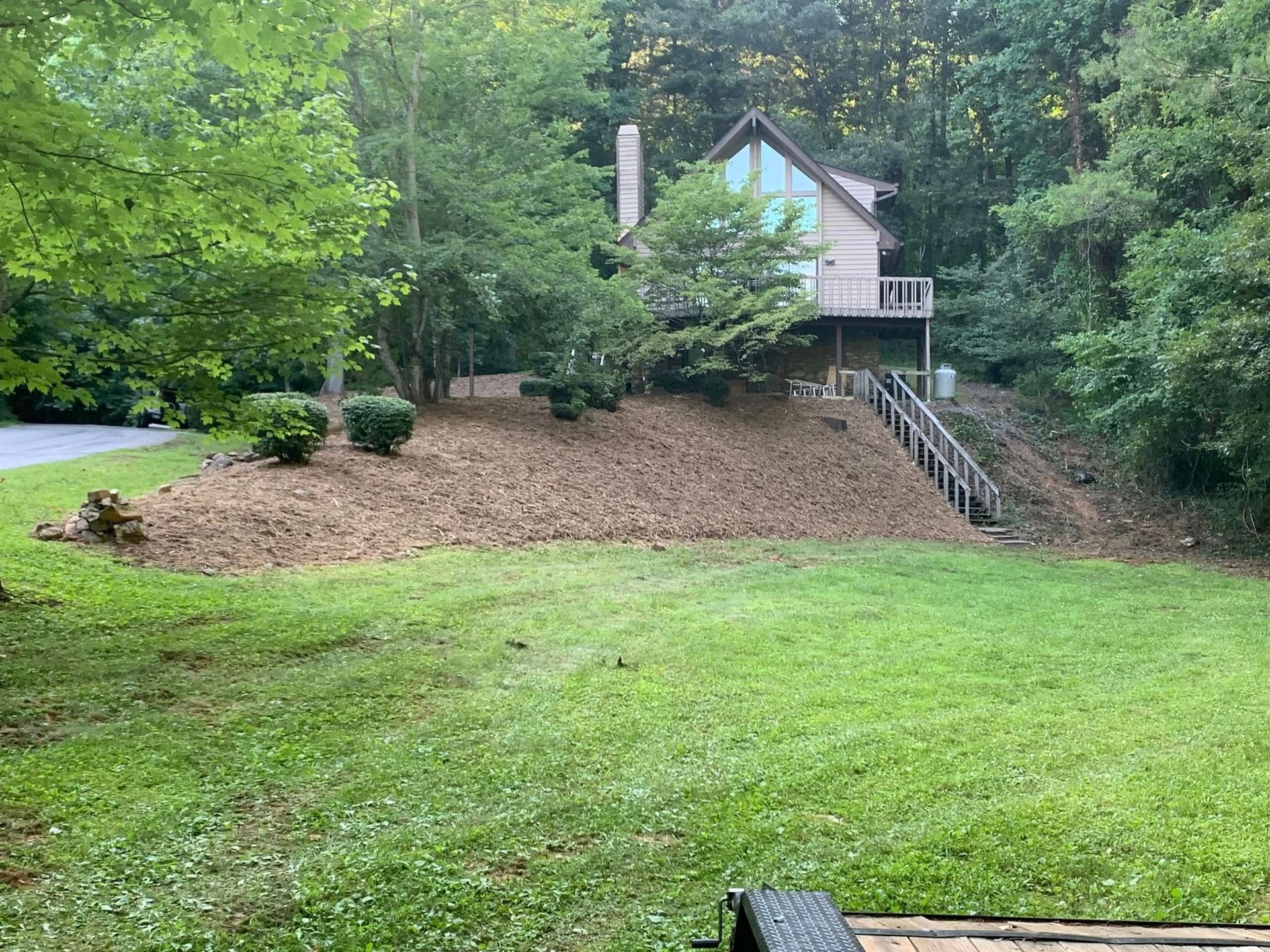 A two-story house nestled in a lush, green forest sits atop a gently sloping, mulched hill with a wooden staircase.