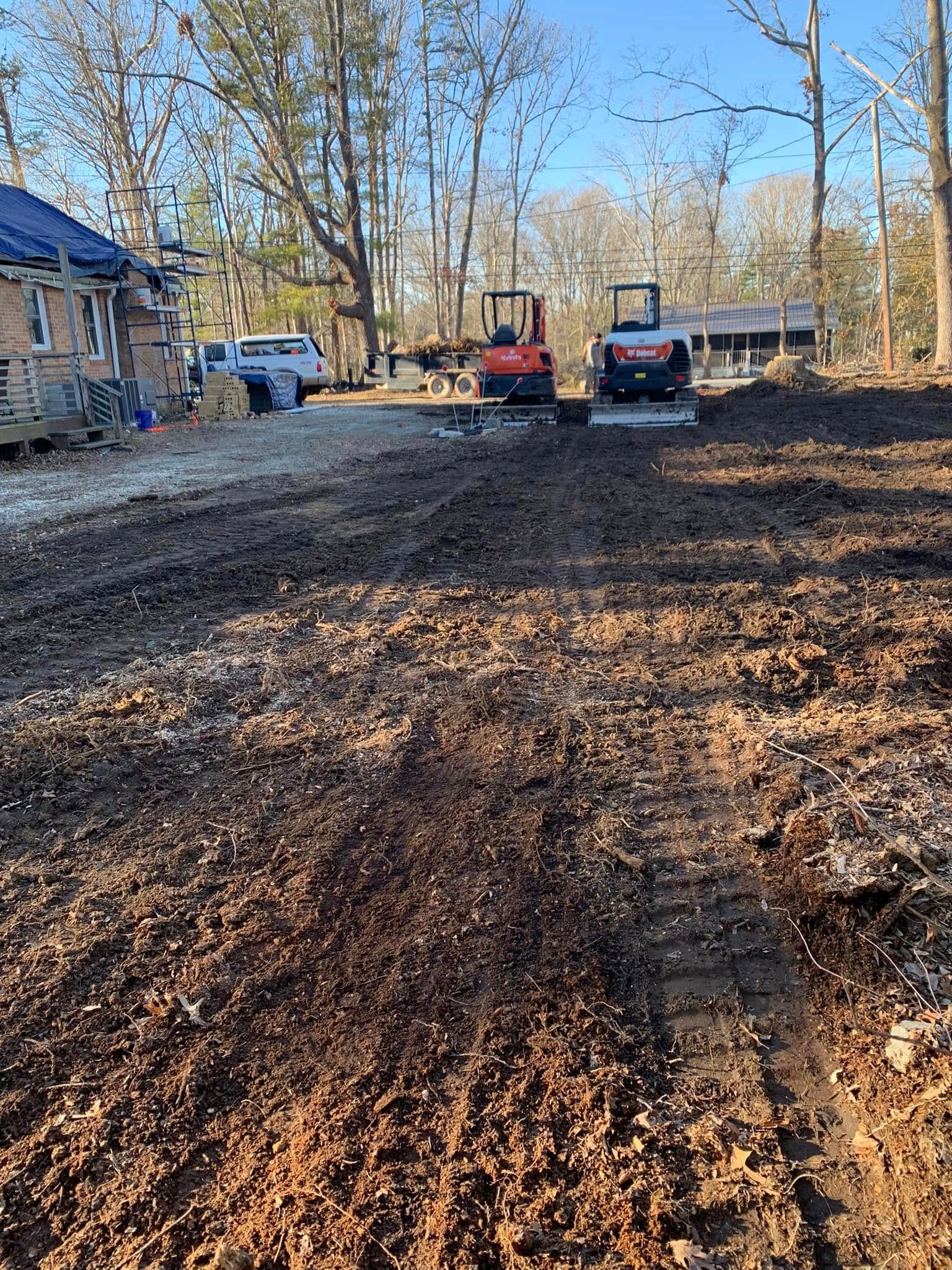 Two construction vehicles sit on a muddy, cleared plot of land near a house and trees under a clear blue sky.