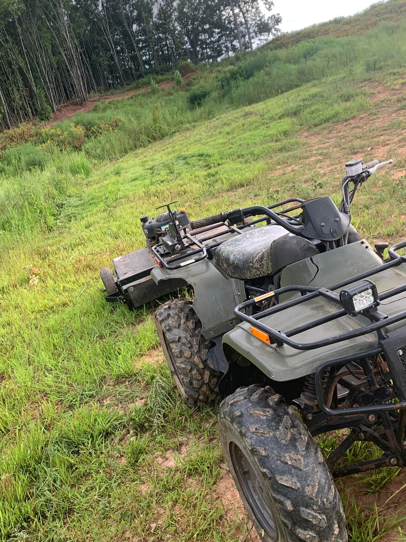 An olive green ATV with a pull-behind mower attachment sits in a grassy, sloped field near a wooded area.