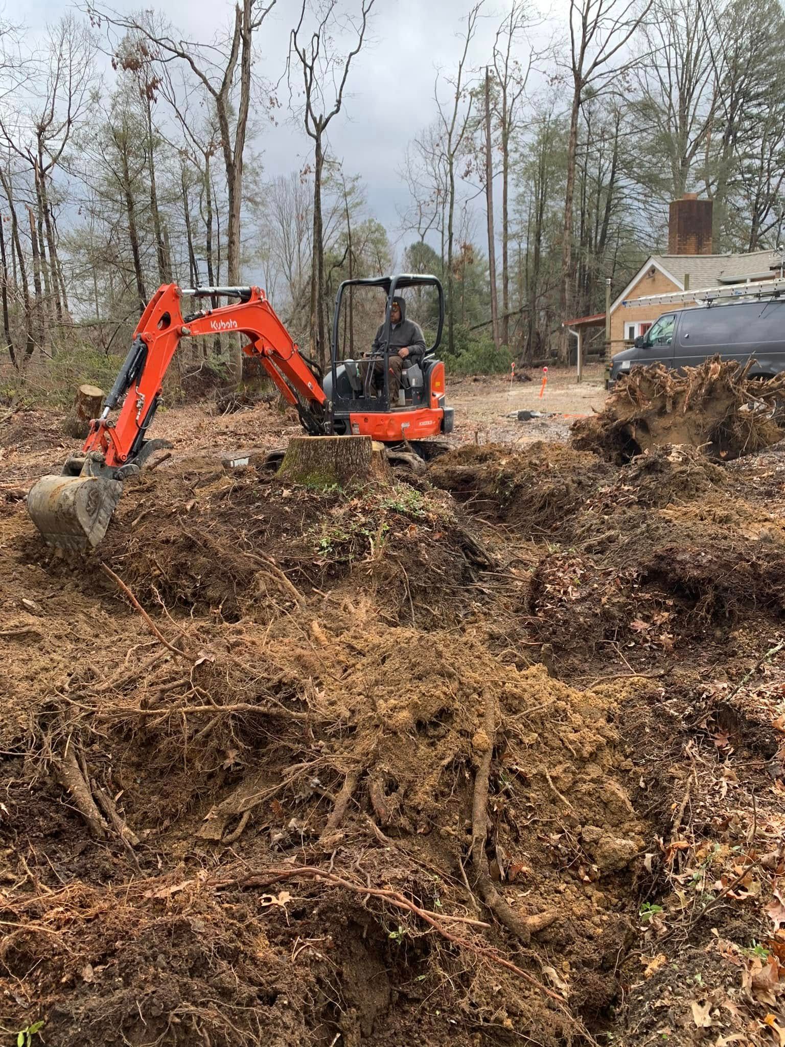 An orange mini excavator clearing tree stumps and debris in a wooded residential yard.