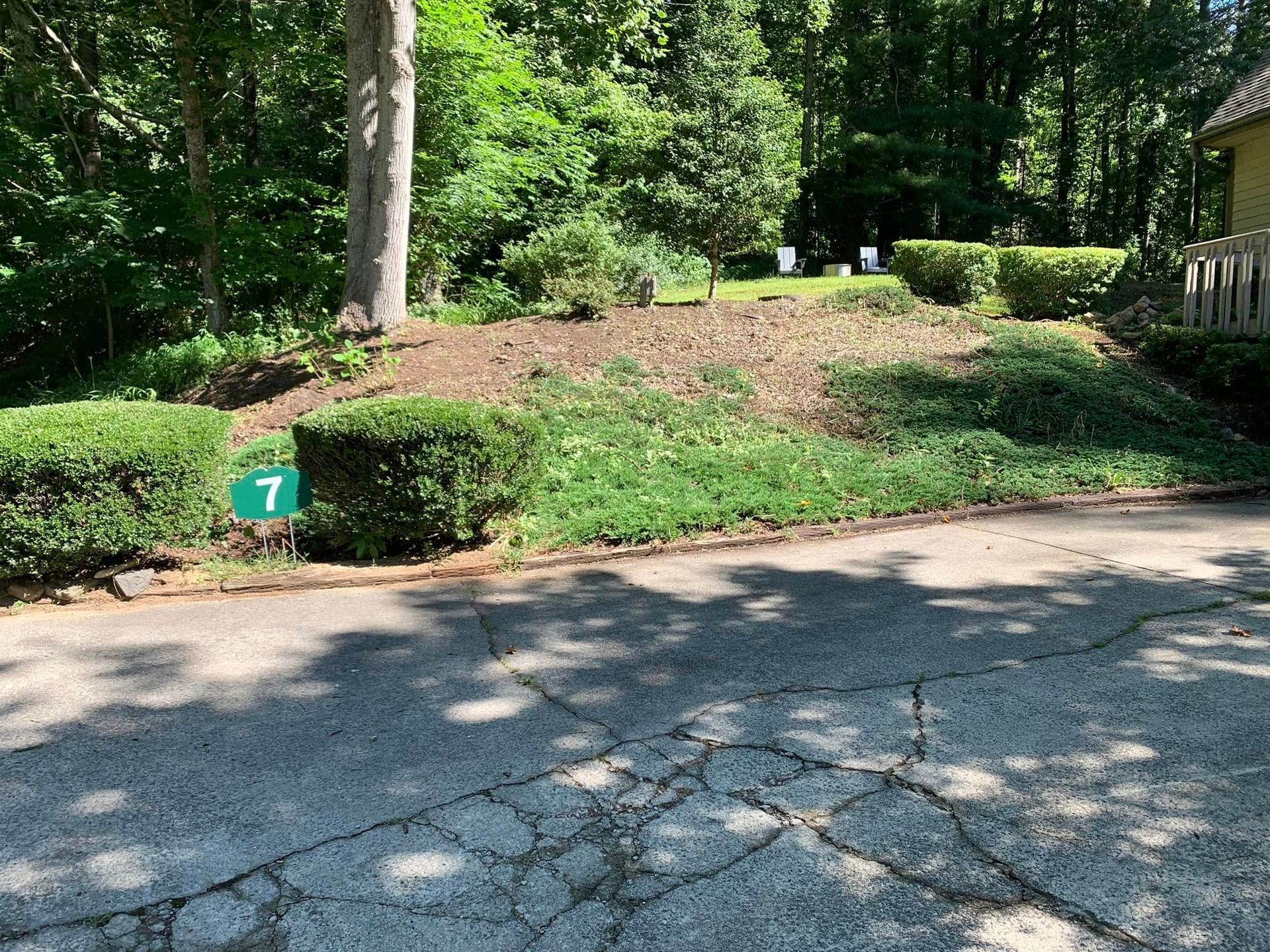 A cracked asphalt driveway leads to a sloping yard with small green bushes and a house number sign marked 