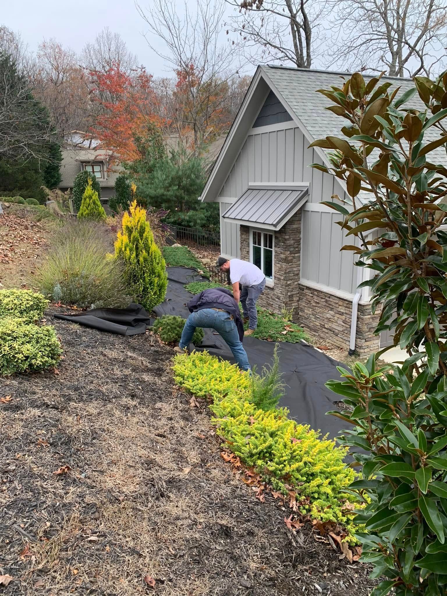 Two people install landscape fabric in a garden bed next to a house with grey siding and stone accents.