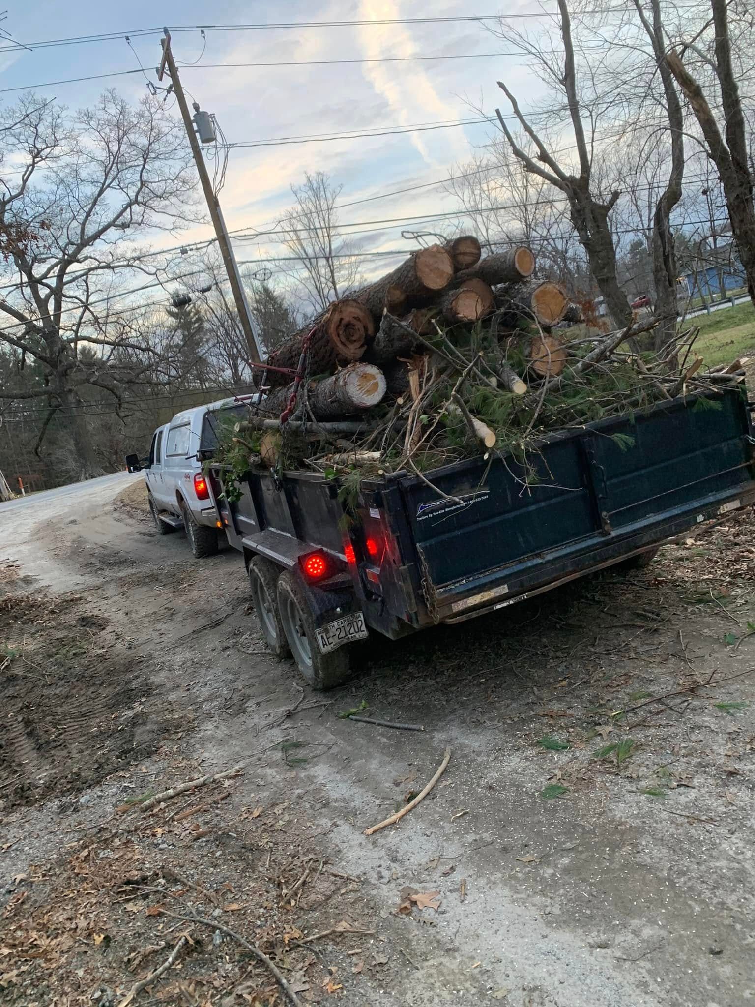 A dark-colored utility trailer, piled high with logs and tree branches, is hitched to a white truck on a dirt road.