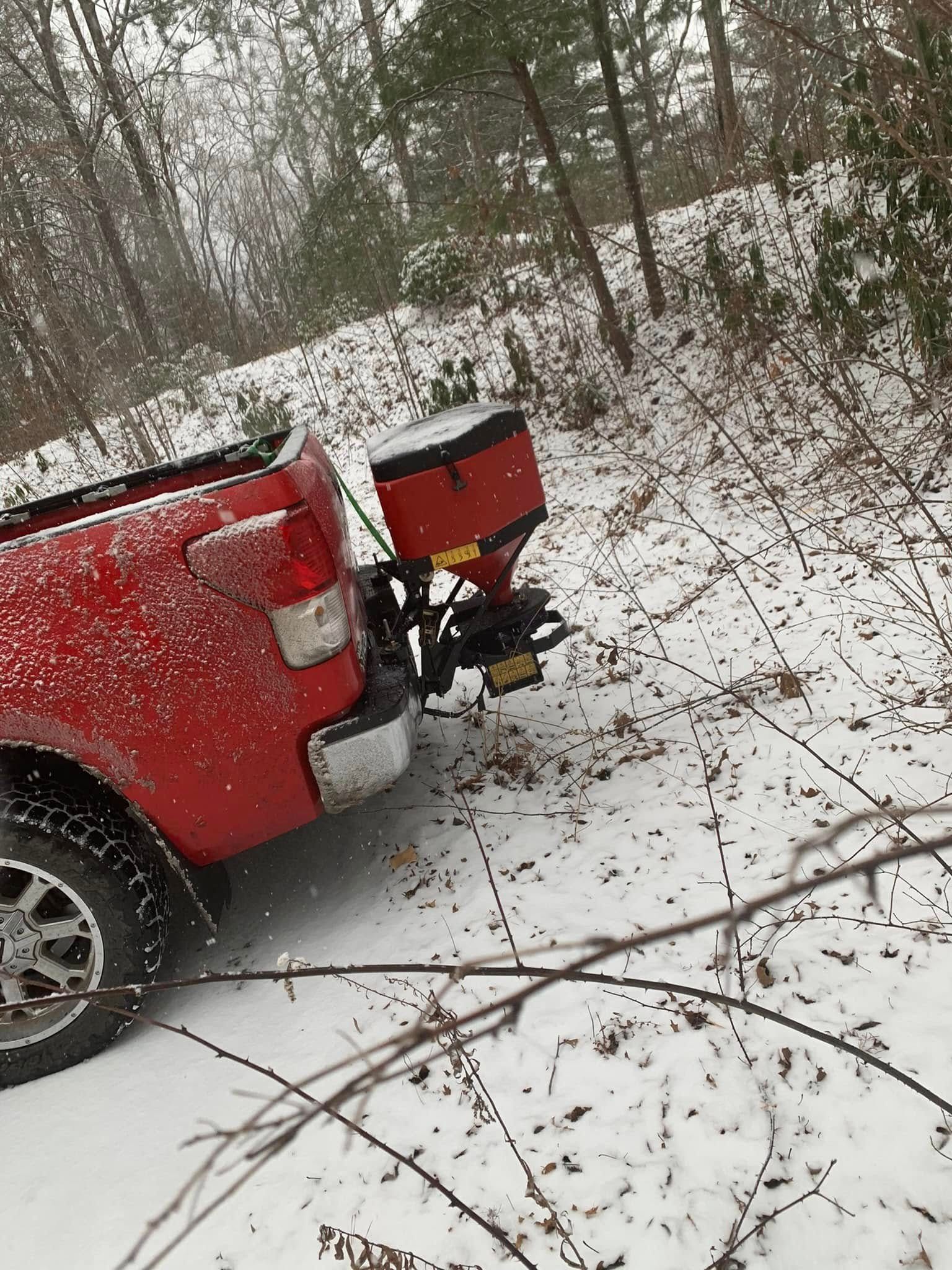 A red pickup truck with a salt spreader attached to its rear hitch, parked in a snowy, wooded area during a light snowfall.