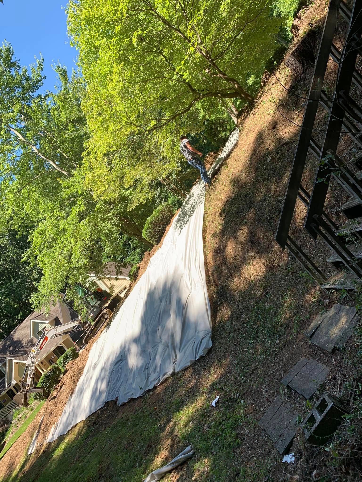 A sloped residential yard featuring a large white tarp covering a section of the hill, surrounded by trees and mulch.