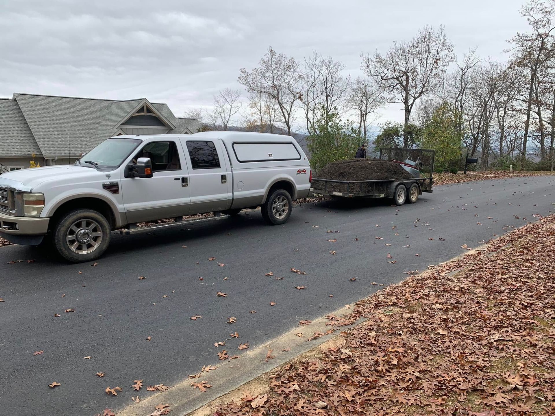 A white pickup truck with a camper shell, parked on an asphalt road, pulling a trailer filled with yard waste.
