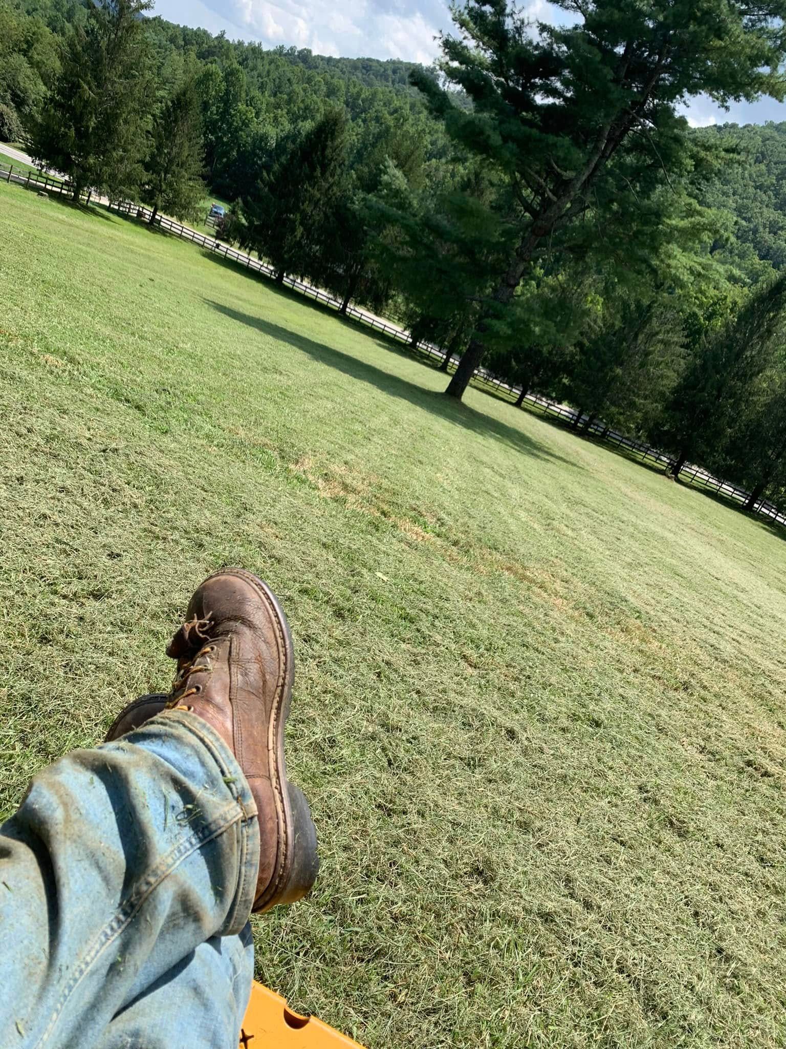 A person wearing blue jeans and a brown leather boot rests their leg against a backdrop of a green field and trees.