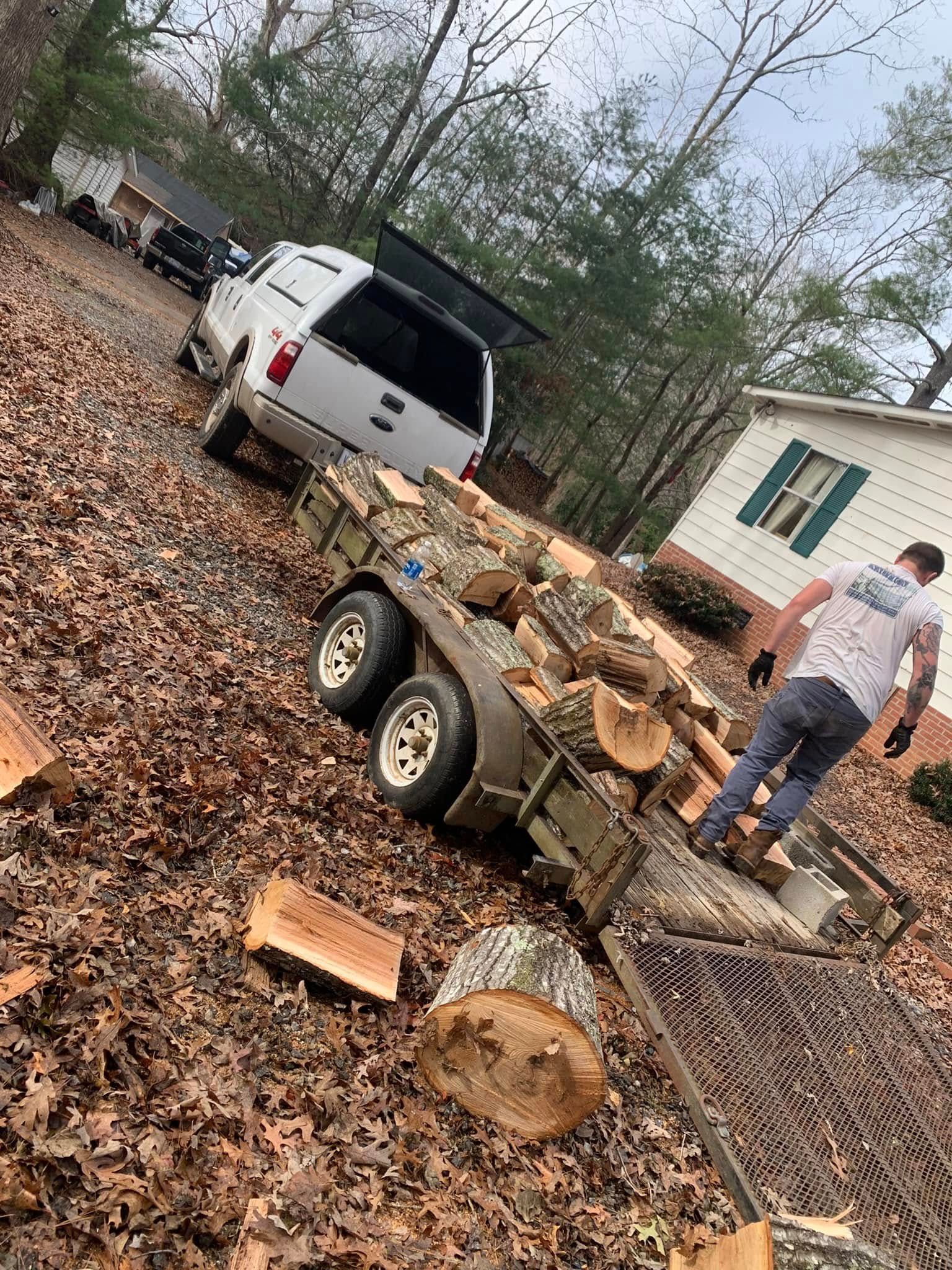 A person loads firewood into a trailer attached to a white truck in a leaf-covered yard.