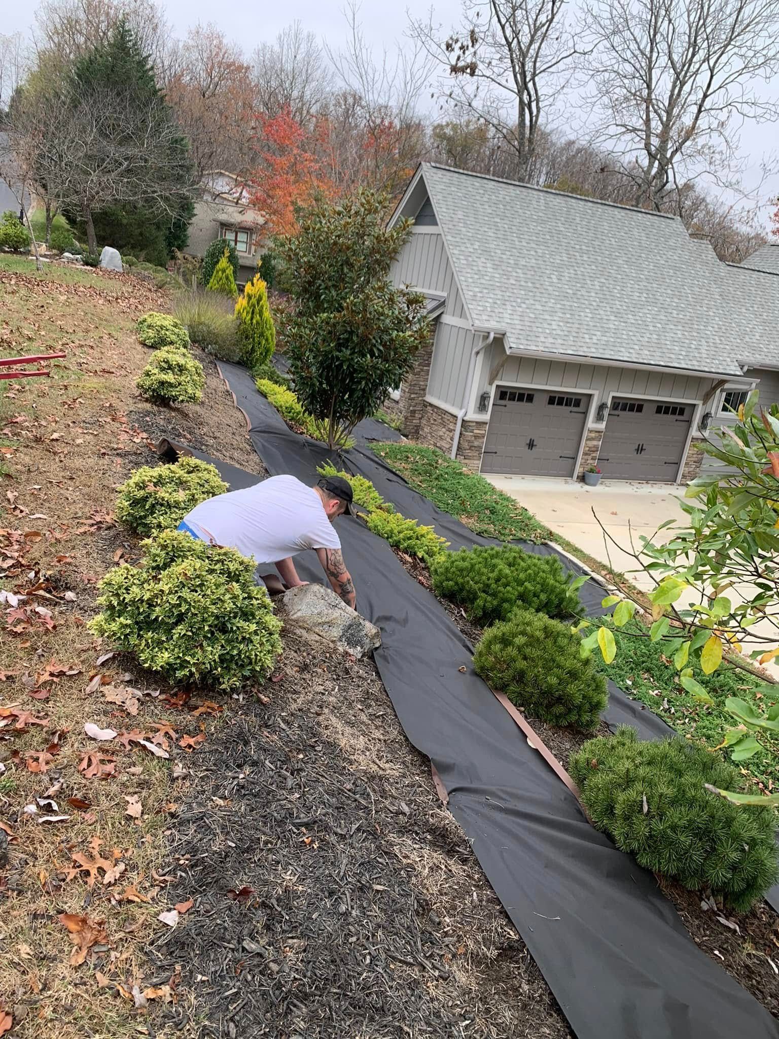A person installs black landscape fabric along a sloped garden bed beside a house with a multi-car garage.