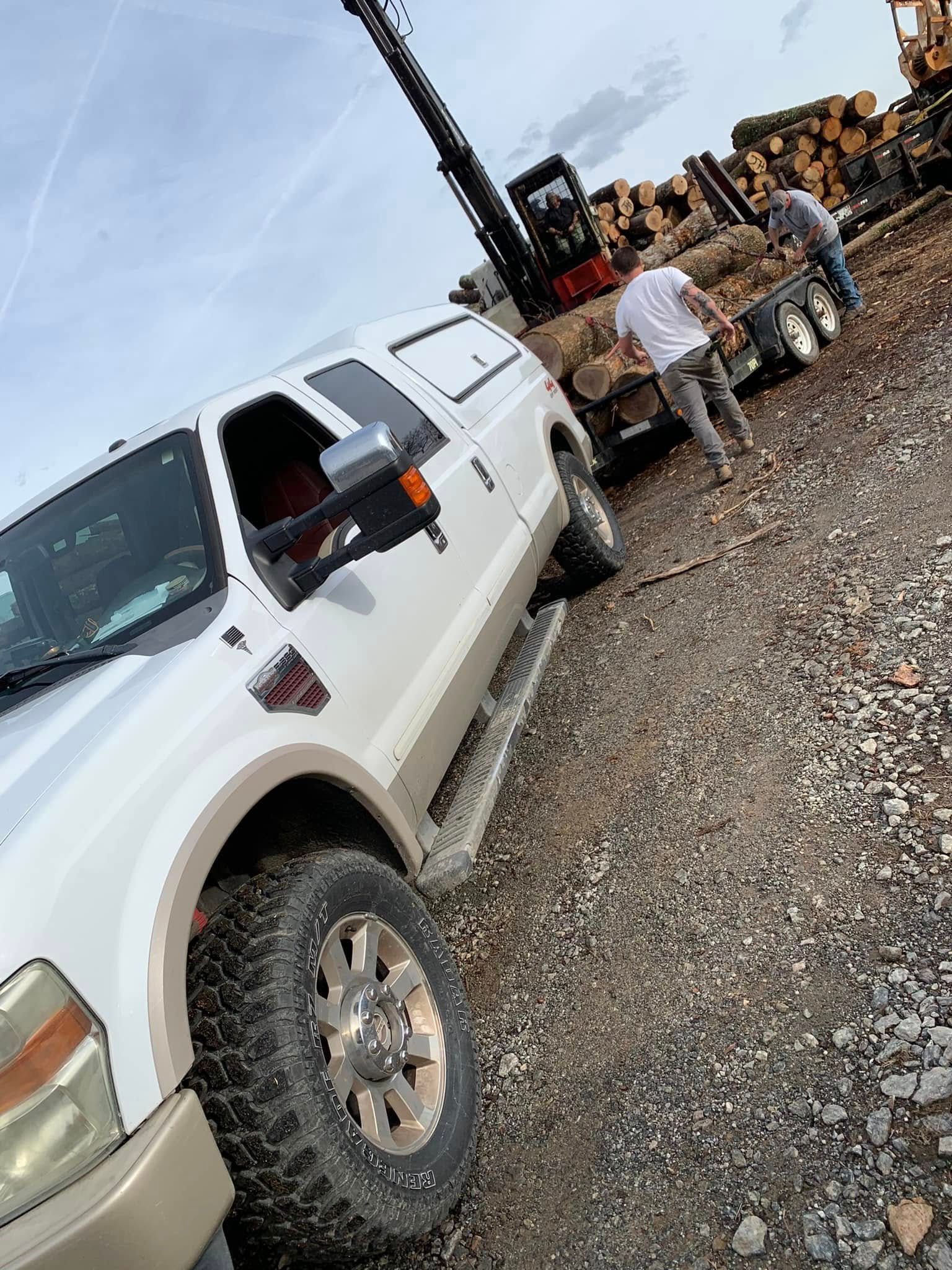 A white truck is parked on a gravel lot as people load logs onto a flatbed trailer using a piece of heavy machinery.